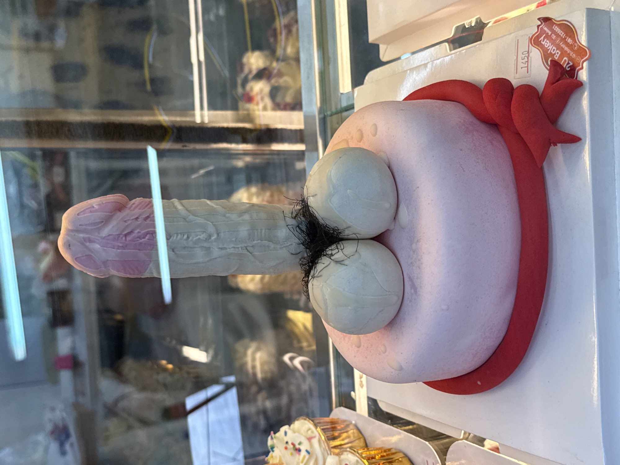 This is a close-up, vertically oriented photograph of a novelty cake shaped like a penis. The cake is displayed behind glass, likely in a bakery case, given the presence of other pastries in the background and a sticker with "2U Bakery" and a phone number visible on a white box behind the cake. The penis cake is sculpted with detail, featuring a pink-tipped head and a white shaft with ridged texture. Two spherical shapes, resembling testicles, are attached to the base of the shaft, with dark, fuzzy material representing pubic hair. The cake is also decorated with a red ribbon or band encircling the base of the testicles and extending outwards. The lighting suggests an indoor environment, possibly a shop or market, with reflections on the glass surface obscuring some of the background details. The time of day is not discernible, but the setting appears to be a commercial establishment. The overall scene is a display of a humorous or provocative cake in a retail setting.