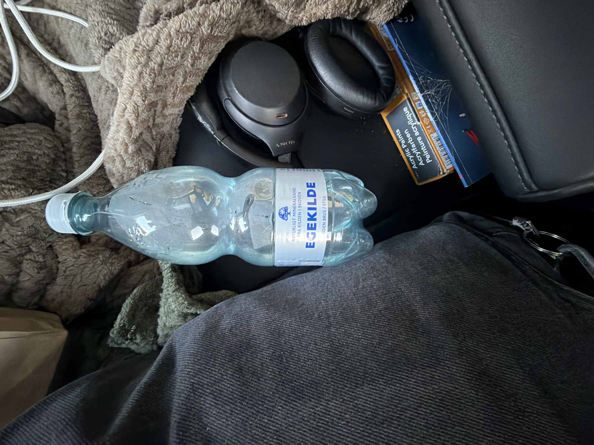 The image shows a close-up, overhead view of personal items resting on what appears to be car seats or a similar upholstered surface. In the center of the frame is a clear plastic bottle of "EGEKILDE" mineral water, with a blue and white label. To the right of the water bottle are a pair of black Sony headphones. Below the headphones and partially obscured by them is a small box labeled "Acrylic Paints," with multilingual text including "Acrylpaints" and "Peinture acrylique." A brown, fuzzy blanket or throw is draped over the top and right side of the scene, and a section of dark, textured fabric, possibly denim jeans, is visible on the left. The lighting suggests an indoor or vehicle setting. The scene is static, with no observable actions or interactions. The location in the city of Ödsmål, Sweden, is not discernible from the visual elements present in the image.