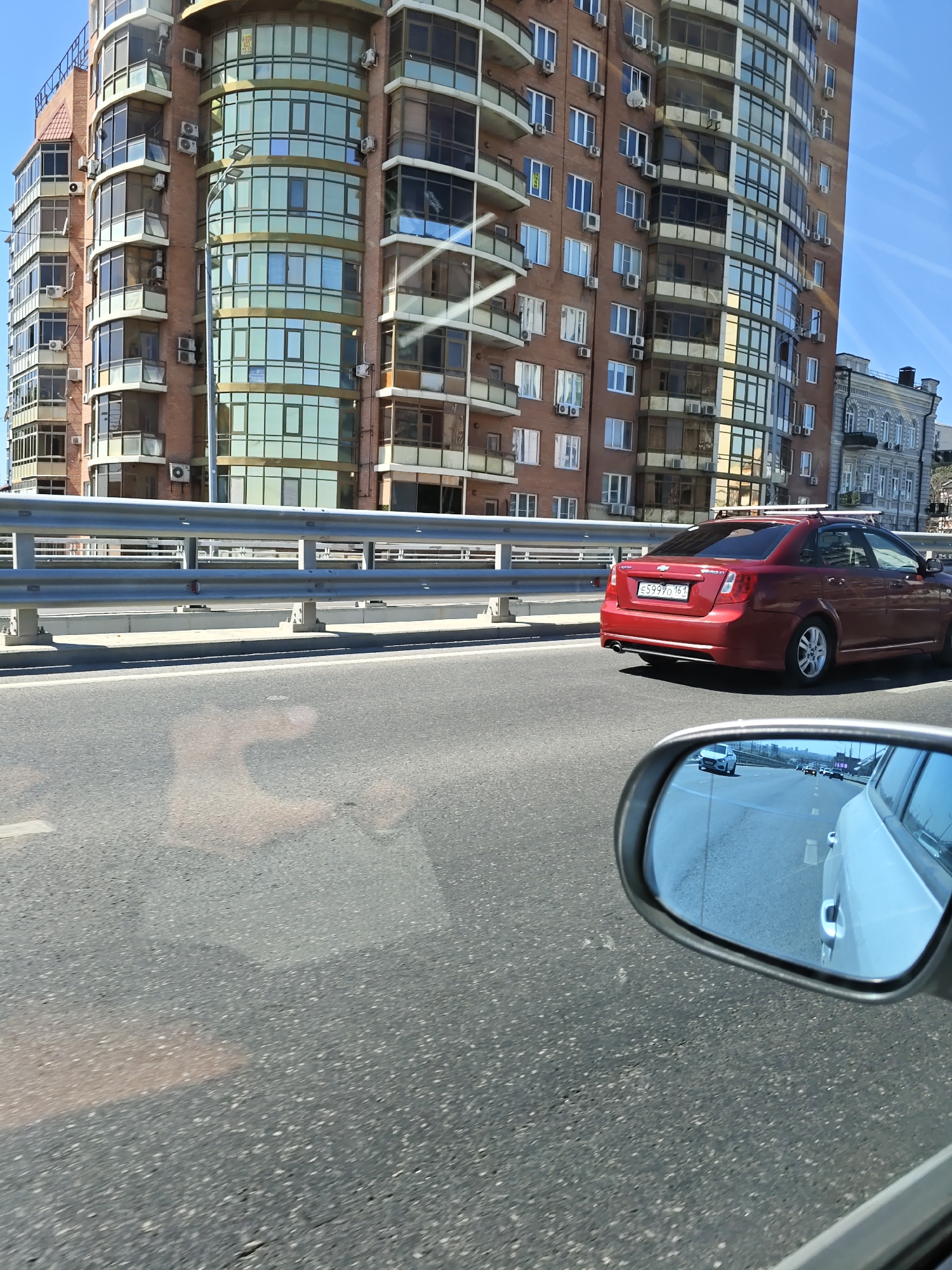 The image depicts a view from a moving vehicle on a sunny day in Rostov-on-Don, Russia. In the foreground, the asphalt road is visible, with a metallic guardrail running along the side of the road, separating it from the sidewalk and a building. The building is a modern, multi-story residential complex with a curved facade, large glass windows, and balconies. Numerous air conditioning units are mounted on the exterior walls of the building. A red Chevrolet sedan is driving on the road, with its license plate clearly visible. In the right side of the image, a car's side mirror reflects other vehicles on the road and a glimpse of the city skyline, suggesting the presence of other cars and buildings in the vicinity. The sky is clear blue, indicating good weather conditions. The scene suggests an urban environment with typical city traffic and architecture.