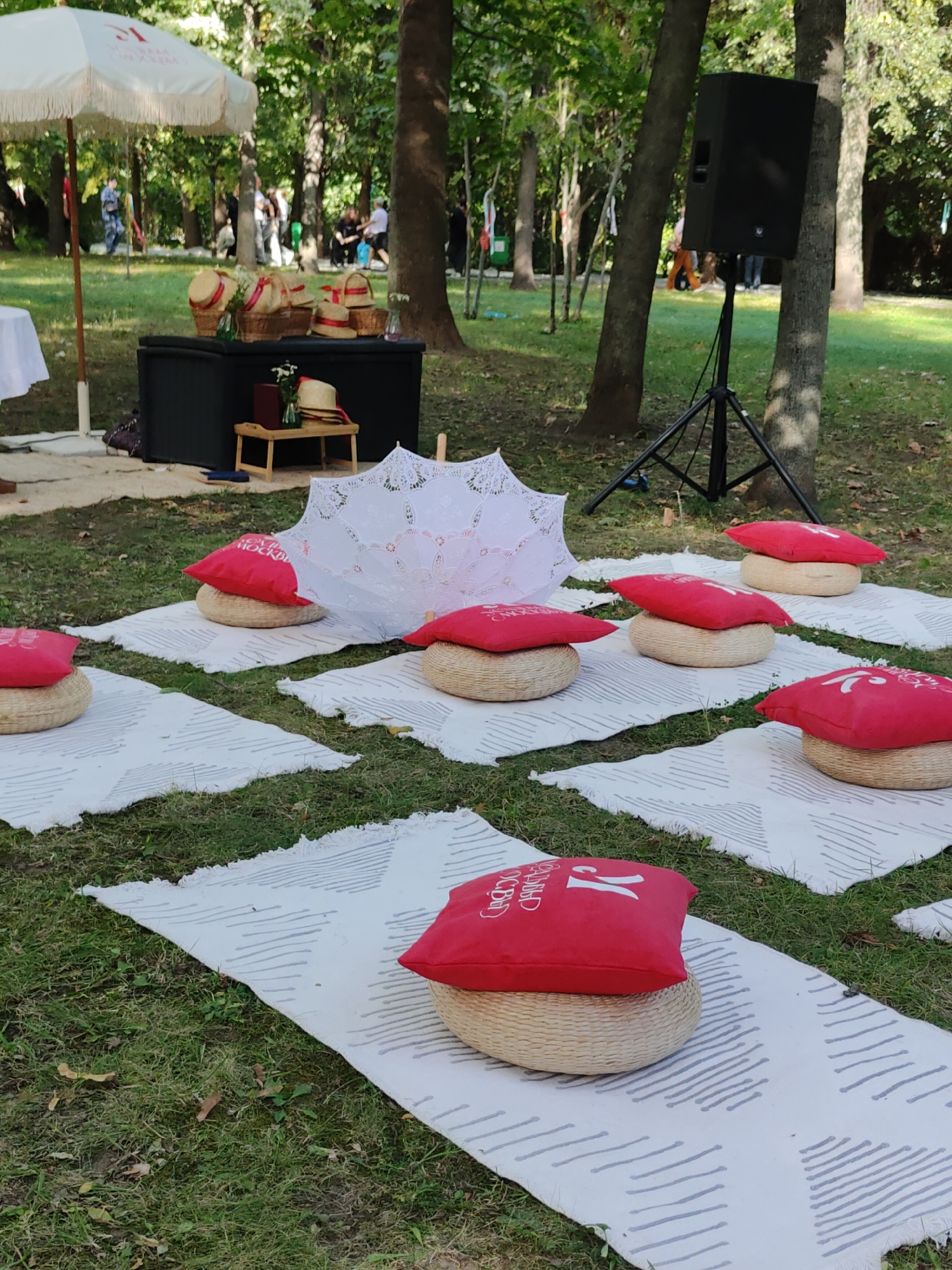 This is an outdoor scene in a park with trees and grass. In the foreground, several white rugs with grey striped patterns are laid out on the grass. On top of these rugs are round woven cushions topped with red square pillows. Each red pillow has a white design on it. To the left of the rugs, a white umbrella with fringe is set up. Behind the umbrella is a dark rectangular table with baskets of straw hats on top. Further back, a black speaker is mounted on a tripod. Several people are visible in the background, some standing and some sitting. The overall setting suggests an outdoor gathering or event.