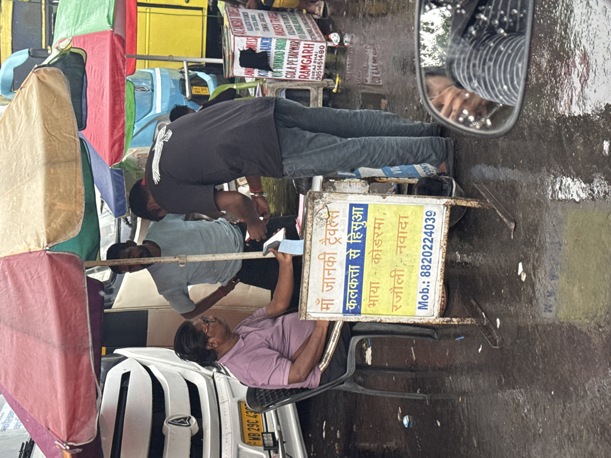 The image depicts a busy urban scene in Kolkata, India, likely during or after a rain shower.
People: There are at least three men visible. One is seated on a chair, wearing glasses and a purple shirt, and looking at something in his hand. Another man is standing behind him, wearing a light blue shirt and observing the scene. A third man is standing to the side, wearing a dark grey shirt and jeans, seemingly interacting with the environment.
Objects: Several objects are present, including a white vehicle with visible license plate "WB 29C 43", a colorful makeshift stall with multiple awnings, and a sign advertising "Maa Janki Travels" with routes to "Hishua via Koderma, Rajauli, Nawada", and a mobile number "8820224039". There is also a sign advertising "Gola-Petarwar Ramgarh, Suraj Bhai 9051451480"
Environment: The setting is a city street, likely after rain, as the ground is wet and reflective. The composition is somewhat chaotic, suggesting a bustling commercial area.