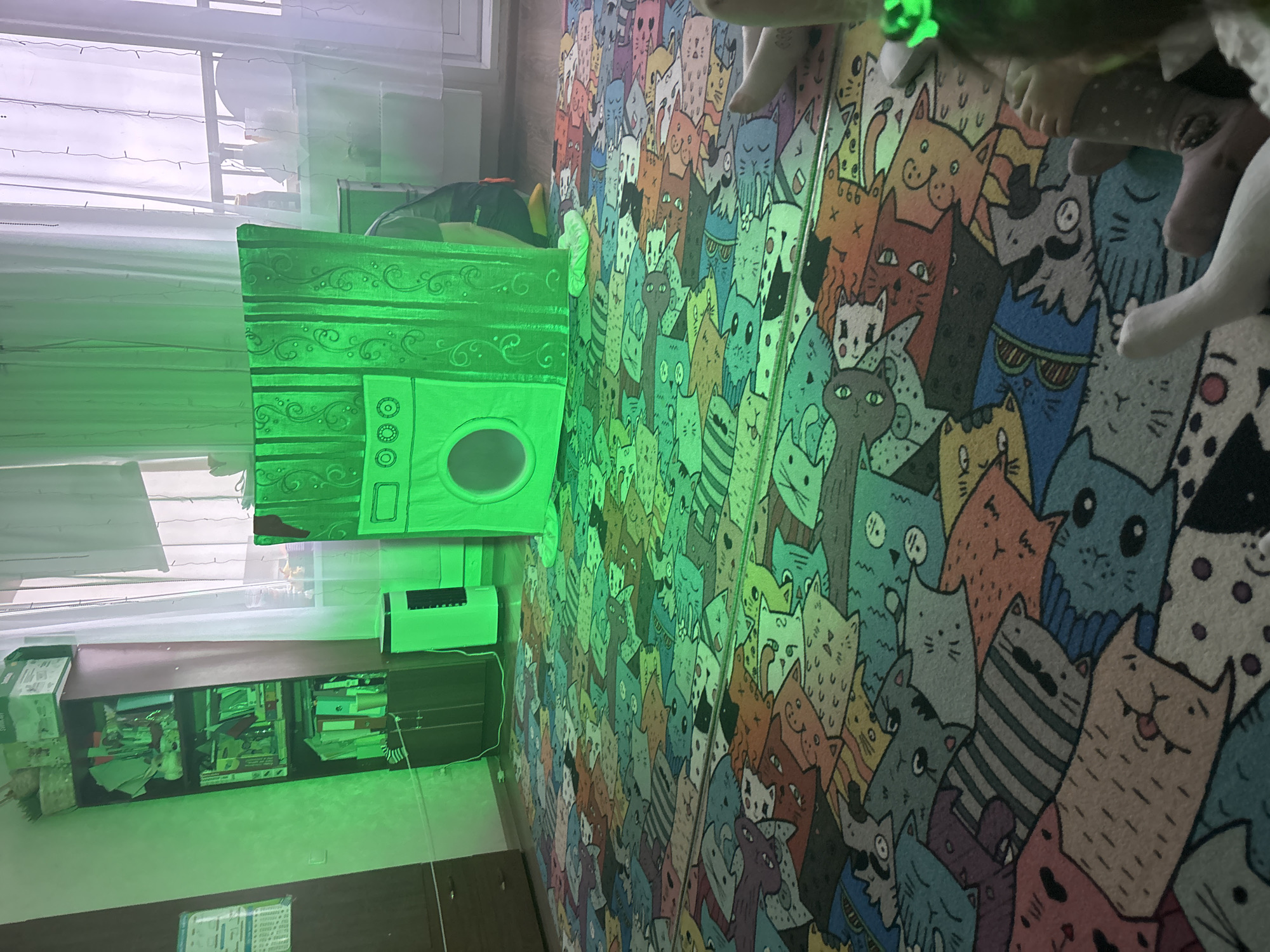 The image displays the interior of a room, likely a bedroom or play area, dominated by a large, colorful rug covering the floor, intricately patterned with numerous cartoon cat faces in various hues including orange, blue, green, grey, and black. The room is bathed in a distinct green ambient light emanating from an internal source.
Against the left wall, a bright green illuminated object, resembling a decorative toy washing machine with ornate scrollwork and a circular opening, is situated on a surface. Above this object, a window fitted with translucent white horizontal blinds is visible, adorned with delicate fairy lights. Below the green object, a white electronic device, possibly an air conditioner or fan, is mounted on the wall. Further down the wall, a dark wooden shelf unit holds an assortment of books, folders, and various miscellaneous items. A white rectangular sign with illegible text is affixed to the wall beneath the shelf unit.
Several small plush toys, one notably emitting a green glow, are scattered on the floor towards the foreground. No people are visible, and no specific activity is taking place; the scene is a static interior view. The time of day is ambiguous; while some daylight filters through the window, the pervasive green artificial illumination creates a distinctive, whimsical, and potentially evening or low-light atmosphere. The room appears lived-in, with its decorative elements and stored items.