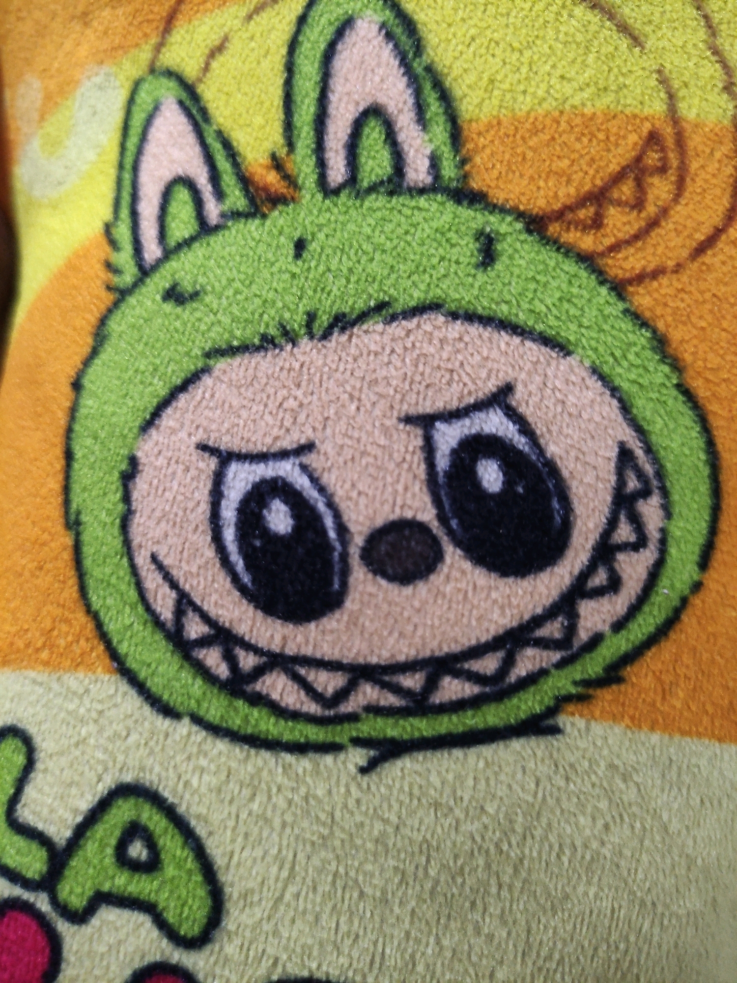 The image is a close-up of a fuzzy, cartoonish character printed on fabric. The character has large, green ears, a round, light brown face with black eyes and a wide, jagged smile. The background is a gradient of yellow and orange. There is text printed on the fabric below the character, but it is partially cut off and illegible. The image does not provide any information about the location in the city of Bulukumba, Indonesia.