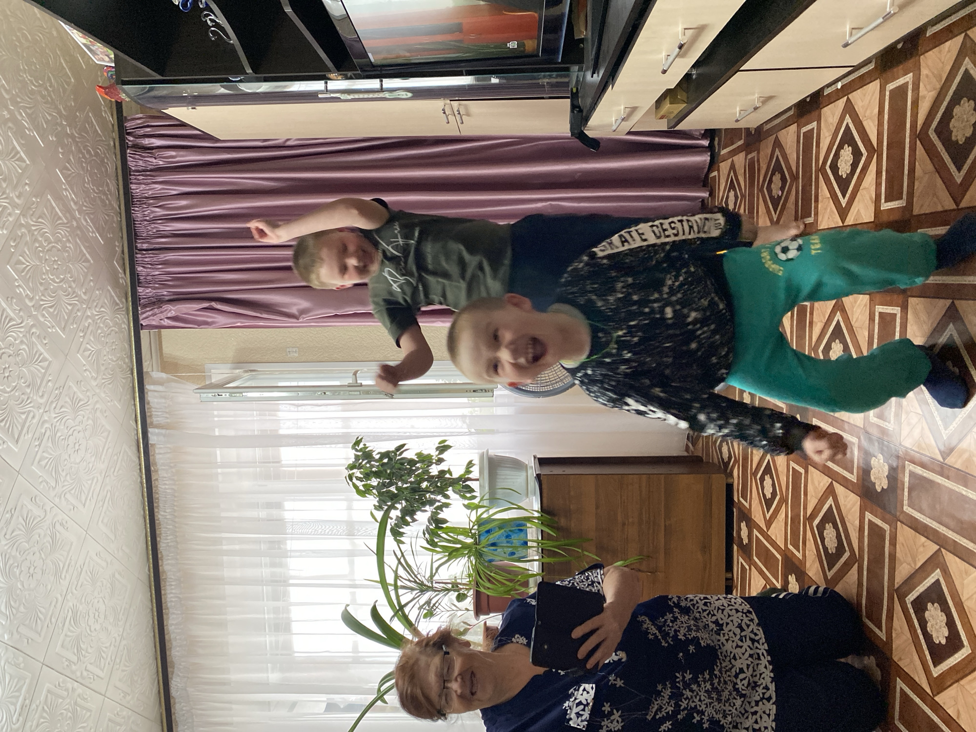 A photograph depicts a scene in Bely Klyuch, Russia. An elderly woman wearing glasses and a patterned blue and white shirt is sitting on the floor and holding a tablet or large smartphone. Two young boys are positioned behind her. One boy is wearing a dark green t-shirt. The other boy wears a patterned shirt and green pants. A large potted plant stands to the left of the woman, positioned in front of a window covered with white sheer curtains. A brown cabinet sits behind the woman. The floor is tiled with brown and beige patterns. Above the boys, a purple curtain is visible below what appears to be a television and storage units mounted on the wall. The interior space has patterned white wallpaper.