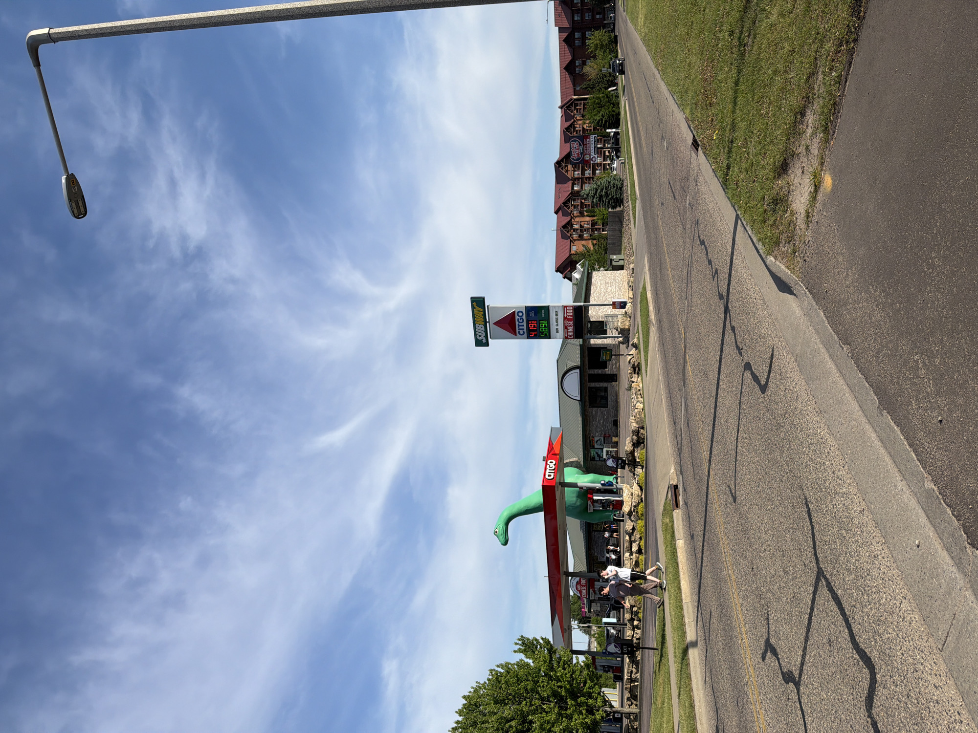 The image depicts a street scene in Lake Delton, United States of America, showing a Citgo gas station with a large, green dinosaur statue next to it. The gas station has a sign displaying gas prices, and a Subway is co-located there. A couple is near the gas pumps. Beyond the gas station are commercial buildings and a tree line. A light pole is visible on the left side of the image. The sky is blue with scattered clouds. The street has visible cracks and repairs, with a grassy shoulder.