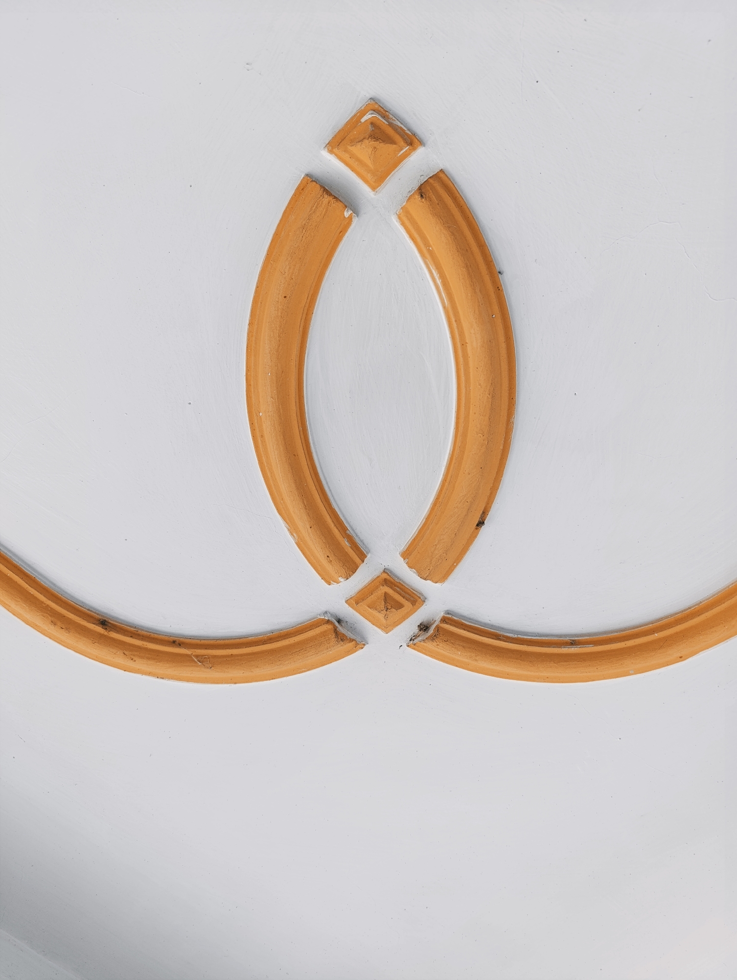 This is a close-up, eye-level, vertical, full-shot photograph of an ornamental molding on a white textured wall. The molding is a decorative element painted in a golden-orange hue. It features a symmetrical design with two elongated oval shapes meeting at the center, forming a bow-tie like appearance. At the top and bottom, the oval shapes are capped by small, diamond-shaped elements, also in golden-orange. The texture of the wall is visible, with subtle imperfections and variations in the paint. The lighting is even and appears to be diffused, casting no harsh shadows. The location is specified as Vashisht, India.