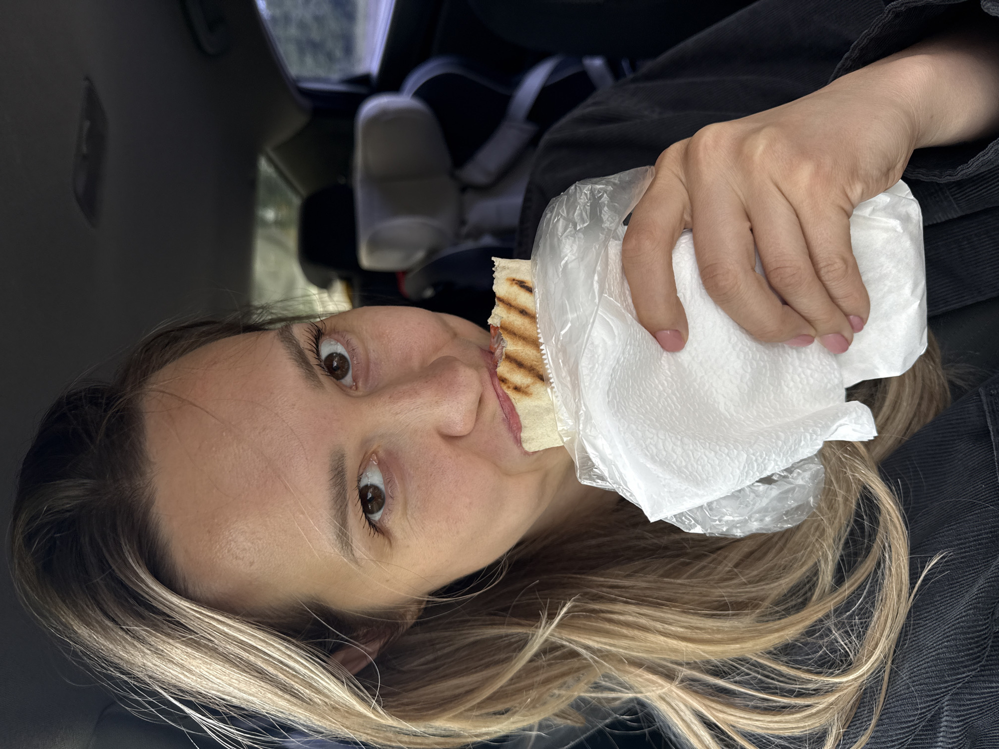 The image captures a close-up of a young woman eating inside a car, rotated 90 degrees counter-clockwise. She has long, light brown hair with blonde highlights framing her face. Her wide-open, dark brown eyes look directly at the viewer, and a slight hint of food is visible near her mouth, suggesting she is mid-bite.
She is holding a grilled wrap or sandwich, which is partially covered by a transparent plastic bag and a white paper napkin. Her right hand, adorned with light pink nail polish, is visible holding the food. She appears to be wearing a dark, possibly corduroy or ribbed, jacket or top.
The background shows the interior of a car, including what appears to be part of a car seat or passenger seat in muted tones, indicating the setting is within a vehicle. The natural light filtering in suggests it is daytime. The woman's expression is one of engaged attention or mild surprise, possibly captured candidly while she was eating. There is no visible text in the image. The location context provided is Chaikovsky, Russia.