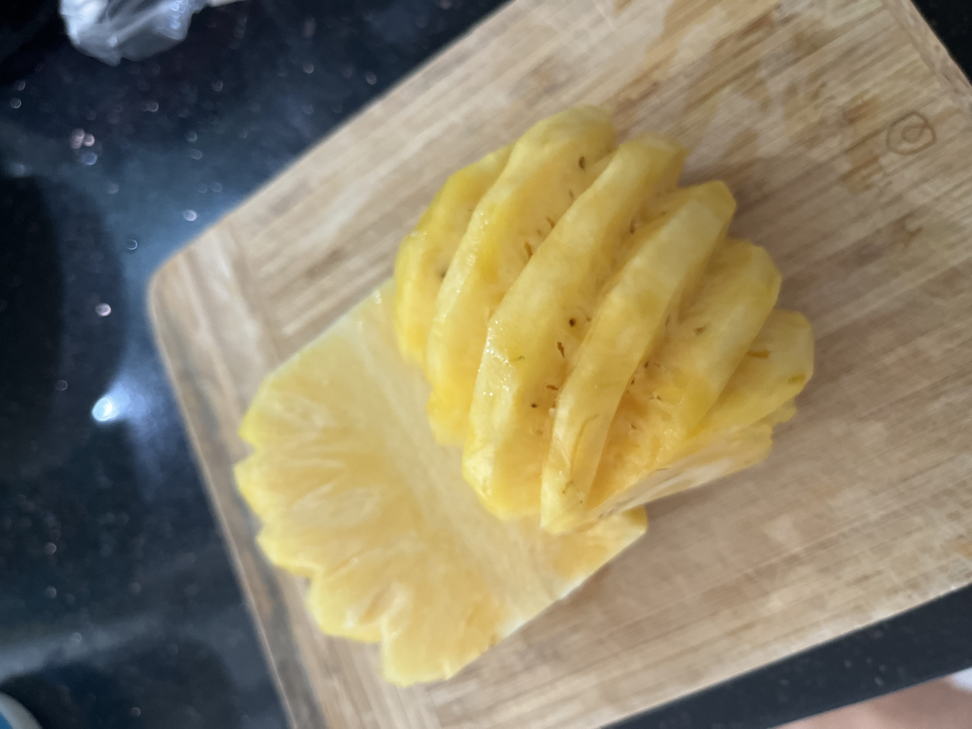 A wooden cutting board holds cut pieces of pineapple. Some pieces are stacked in a spiral arrangement. The surface next to the cutting board is dark and reflective, suggesting a kitchen countertop. The image was taken indoors. It is unknown if the image was taken in Bac Nha Trang Ward, Vietnam.
