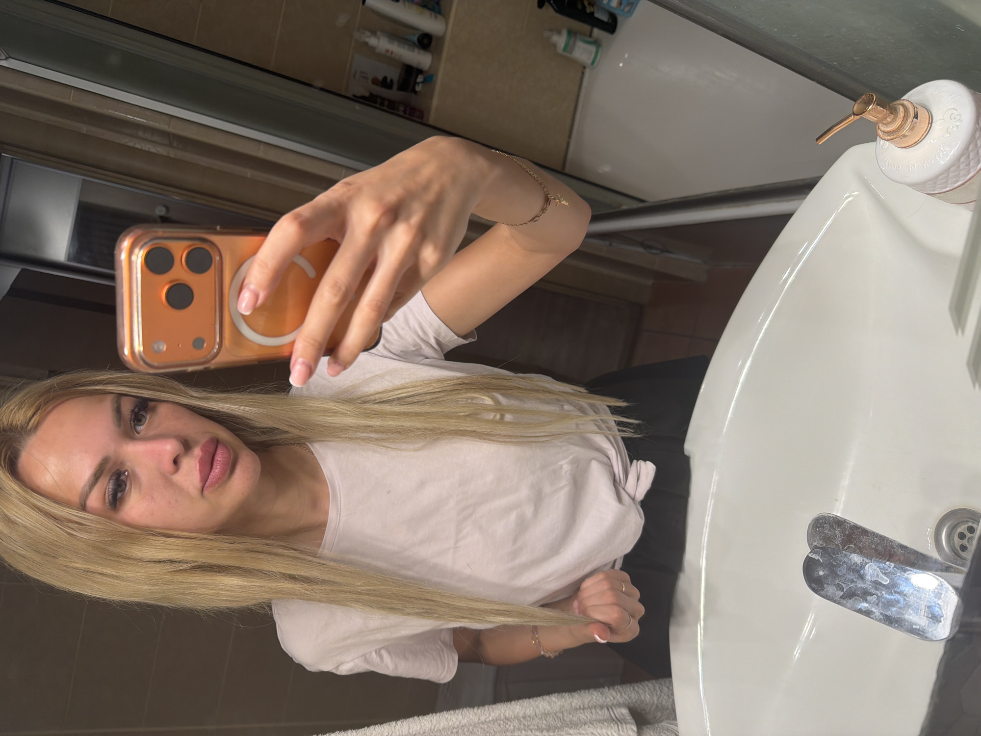 A woman with long blonde hair is taking a mirror selfie in a bathroom. She is wearing a light pink t-shirt tied at the waist and black bottoms. She is holding an orange-cased smartphone in her right hand. The bathroom features a white sink with chrome faucets, and various toiletries are visible on shelves in the background. The image was taken in Kyiv, Ukraine.