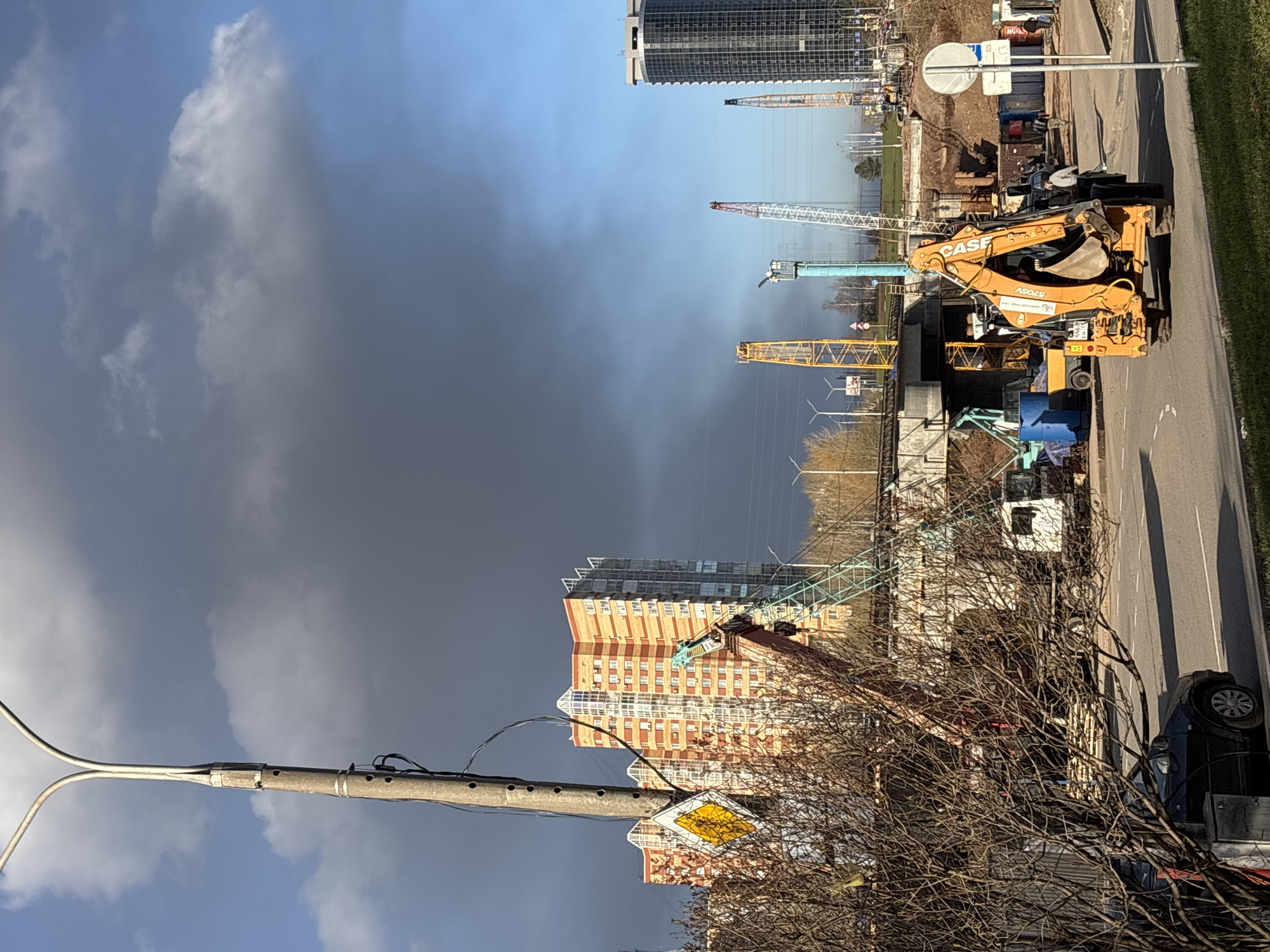 The image, oriented horizontally, captures an active urban and construction scene under a dramatic sky. The left side of the sky is a clear, bright blue, contrasting sharply with the large, dark gray clouds that dominate the right side of the frame, suggesting a changing weather pattern.
In the background, a tall, modern skyscraper with a glass facade stands on the left. To its right, a significant construction site is visible, populated by several large cranes of various colors, including red, white, blue, and yellow, indicating extensive building activity. Further right, a large apartment building with distinctive orange-brown brick-like facades and numerous windows is partially visible behind trees.
The mid-ground features a construction area adjacent to a paved road. A prominent yellow "CASE" excavator, with "AS046" and "STONE" branding, is parked on the edge of the road. A partially visible license plate on the excavator reads "P59 F051". Other smaller construction equipment and materials are scattered around this area.
In the foreground, a gray street pole equipped with wires and a yellow diamond-shaped sign with a black border (likely indicating a main road) stands on the left. A blue and white circular sign, possibly a traffic regulation sign, is also visible. A paved road runs along the right side of the image, with white lane markings. A dark car is parked on the far right, partially obscured by bare tree branches that fill the lower right corner, indicating a cooler season.
The scene is illuminated by daylight, casting distinct shadows. The overall impression is one of ongoing urban development in Perm, Russia, under a striking, dual-weather sky.