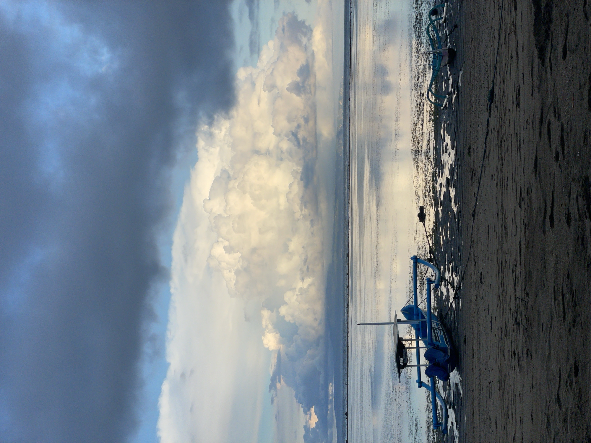 The image depicts a beach scene in Denpasar, Indonesia, under a dramatic sky. A traditional outrigger boat rests on the dark, damp sand, its blue and white frame contrasting with the dark shoreline. The calm water reflects the sky, mirroring a mix of heavy, dark storm clouds and patches of bright white clouds. The division of the sky and the sea creates a balanced composition in the shot.