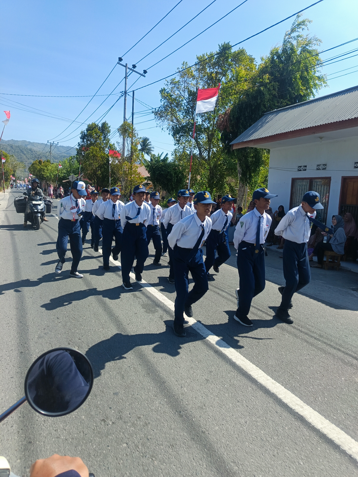 The image depicts a street scene in Tenggela, Indonesia, likely during a parade or procession. In the foreground, a motorcycle rider's perspective is visible, showing a rearview mirror reflecting the rider's dark blue trousers and the road ahead. The main focus is on a group of male students, all wearing white shirts, dark blue trousers, and blue caps, marching in formation. Some of the students are identified with "SMP" or school uniforms. They appear to be in unison, possibly during a school event.
Behind the students, a motorcycle with a rider wearing a dark helmet and a jacket is seen, followed by other participants and onlookers. The Indonesian flag, red and white, is prominently displayed on a flagpole, along with other red and white flags adorning the street. The environment includes a paved road with a white dividing line, trees, utility poles with power lines, and buildings on the right side of the street. The sky is clear and blue, suggesting a sunny day. The background shows a hilly landscape. The overall atmosphere appears to be festive and patriotic.