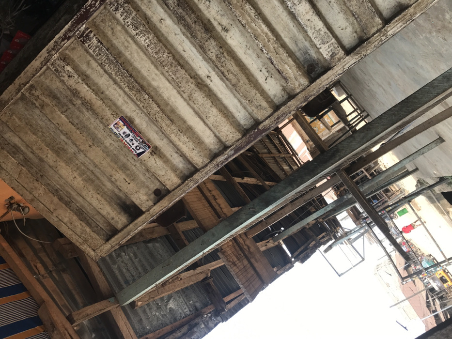 The image depicts an outdoor market scene in Akure, Nigeria, characterized by a network of weathered, makeshift structures viewed from a low, tilted perspective. In the upper right foreground, a corrugated panel, possibly tin or wood, shows significant wear, dirt, and light-colored streaks; affixed to it is a rectangular sticker featuring the words "Bad Spare Parts" and illustrations of vehicle components. Dominant structural elements include multiple rough-hewn wooden beams and at least one prominently featured vertical beam painted a faded light green/teal, all appearing aged and structurally supporting various overhead corrugated metal sheets and wooden planks that form roofs and ceilings.
Through the gaps and beneath these structures, a street or open market area is visible in the mid-ground and background. One individual, wearing a red shirt, is seen walking away from the viewer on what appears to be a dirt or uneven paved road. Further down the street, a yellow tricycle (keke Napep) is partially visible, indicating local transportation. Other distant figures and additional rudimentary market stalls or buildings line the street. The ground immediately beneath the visible structures appears to be concrete or packed earth. The scene is illuminated by natural daylight, with areas under shadow from the overhead coverings.