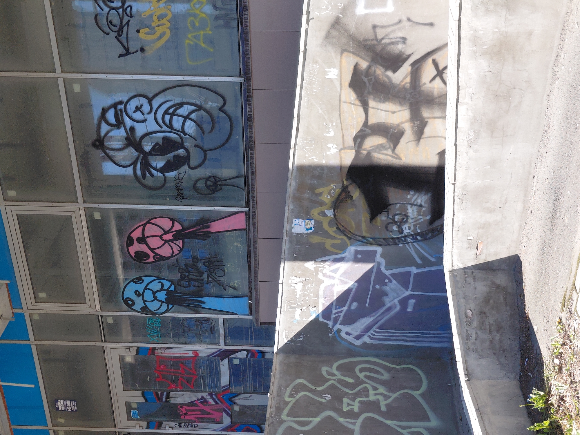 An exterior shot of a building with large windows shows extensive graffiti. The building has a blue awning and a section with a tiled facade. Graffiti covers the windows, featuring cartoonish characters, scribbled tags, and abstract designs in various colors including pink, blue, yellow, red, and black. Below the windows, a concrete ramp or wall is also covered in graffiti. This graffiti includes outlines of figures, abstract shapes, and tags in black and white, with some sections partially obscured by shadows. The ground in front of the structure is paved and shows scattered debris and some green vegetation.