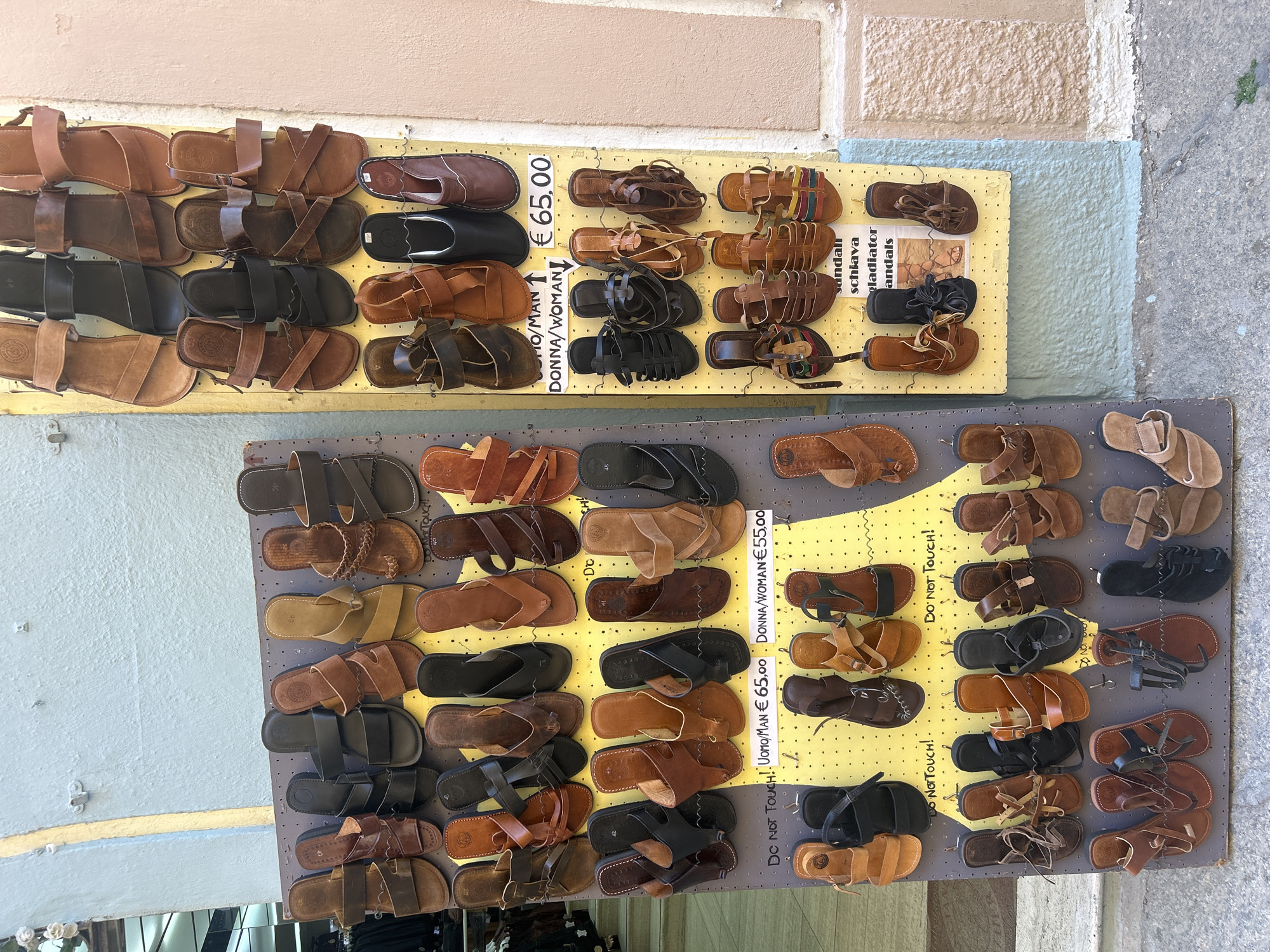 This image shows a display of handmade leather sandals for sale in Alghero, Italy. Two painted wooden boards, one with a yellow background and the other with a gray background, are hanging outdoors. Each board has multiple pairs of sandals in various styles and shades of brown and black. Text on the yellow board indicates prices for men's and women's sandals. Some pairs of sandals are labeled "Do Not Touch!" There are different color schemes of walls in the background, indicating it is somewhere in a street in the city.