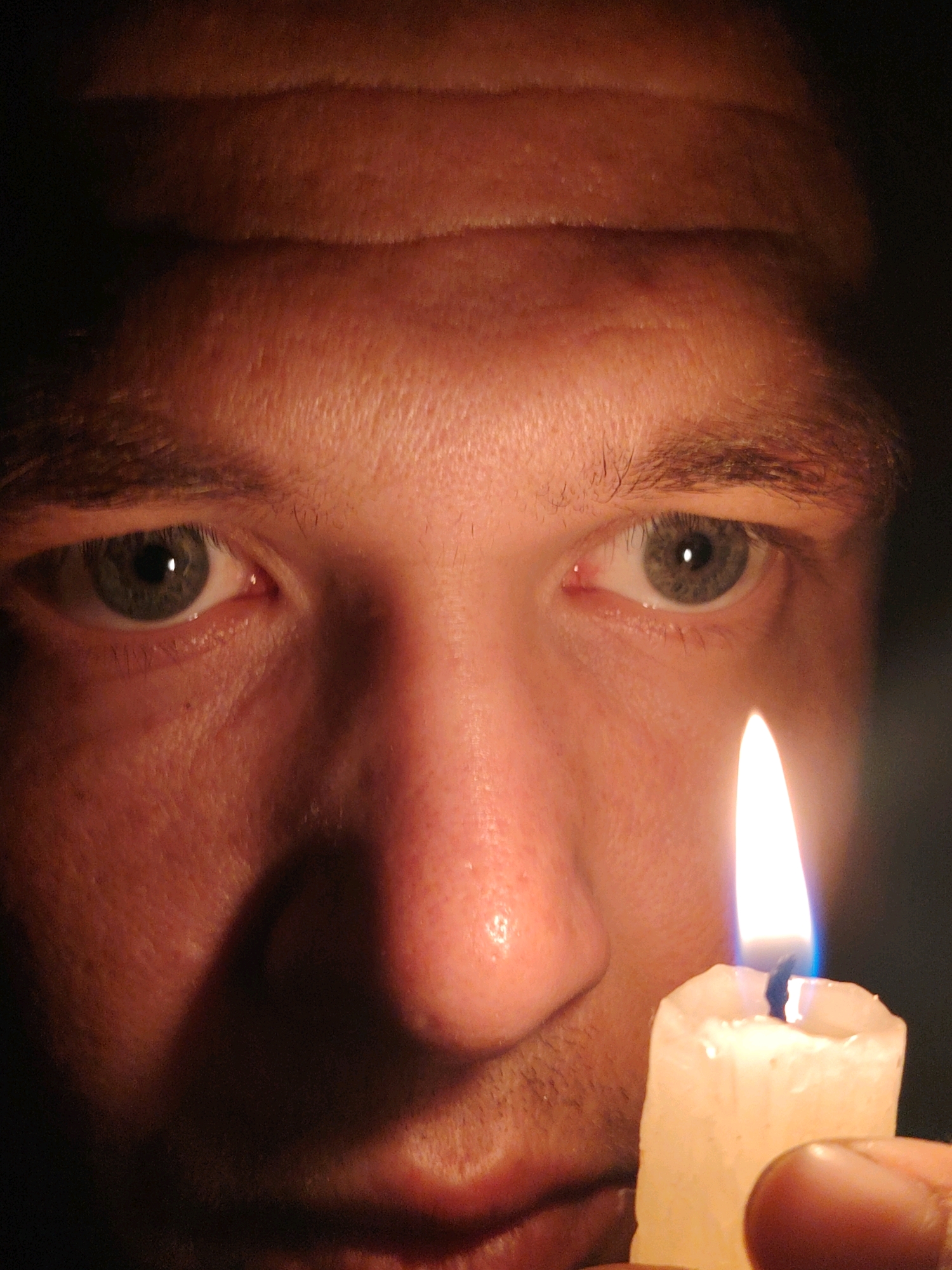 This is a close-up, low-angle, medium shot of a person's face illuminated by candlelight. The person is holding a lit white candle in their right hand, close to their nose and mouth. The flame of the candle is bright white with blue at its base. The person's eyes, which appear to be green, are looking towards the camera. Their forehead is furrowed, and the skin texture is visible. The lighting is dramatic, with strong shadows cast across the face. The background is dark, making the face and candle stand out. There is no identifiable location in Chaikovsky, Russia, or any specific context provided by the image itself.