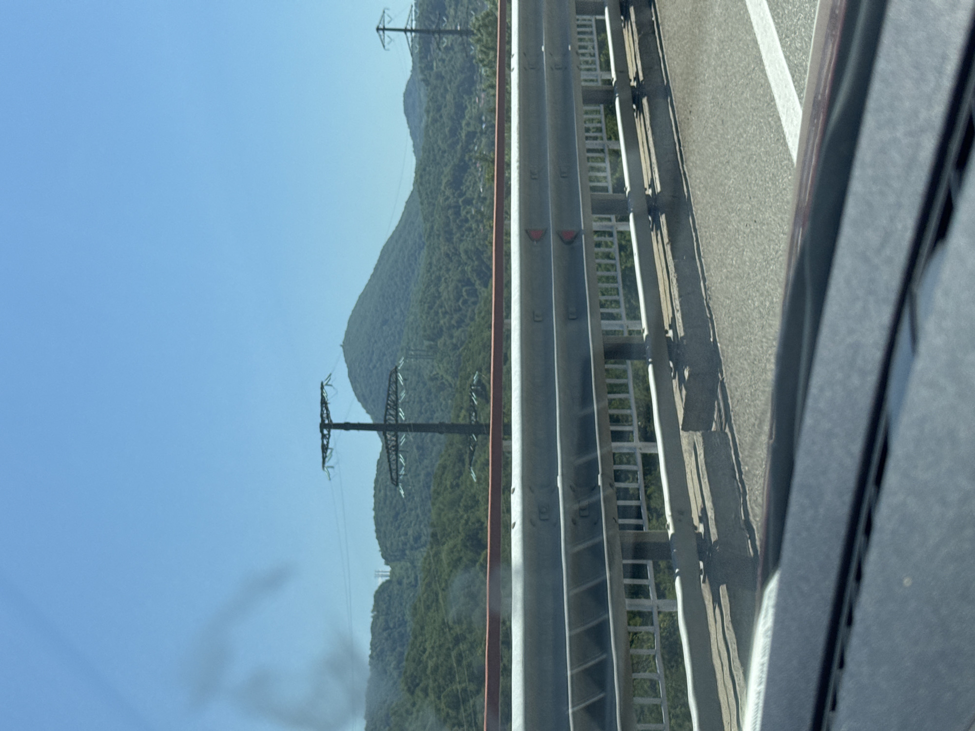 This is a medium-range, eye-level, outdoor, daytime, vertical shot taken from inside a vehicle, looking over a guardrail and towards a mountainous landscape under a clear blue sky. The foreground shows the interior of a vehicle, with a dark dashboard and the edge of the windshield. A grey metal guardrail with a red top rail separates the roadway from a green, tree-covered embankment. Beyond the guardrail, the landscape rises into rolling green hills and mountains. Several power line towers are visible on the slopes, including a prominent, tall one with a wide crossbar and insulators, and another smaller one further in the distance. The scene appears to be a highway in a rural or semi-rural area, possibly in or near Razdolnoye, Russia, given the presence of mountainous terrain and infrastructure like power lines. There is a wispy, dark smudge in the upper left portion of the sky, possibly smoke or a lens flare artifact.