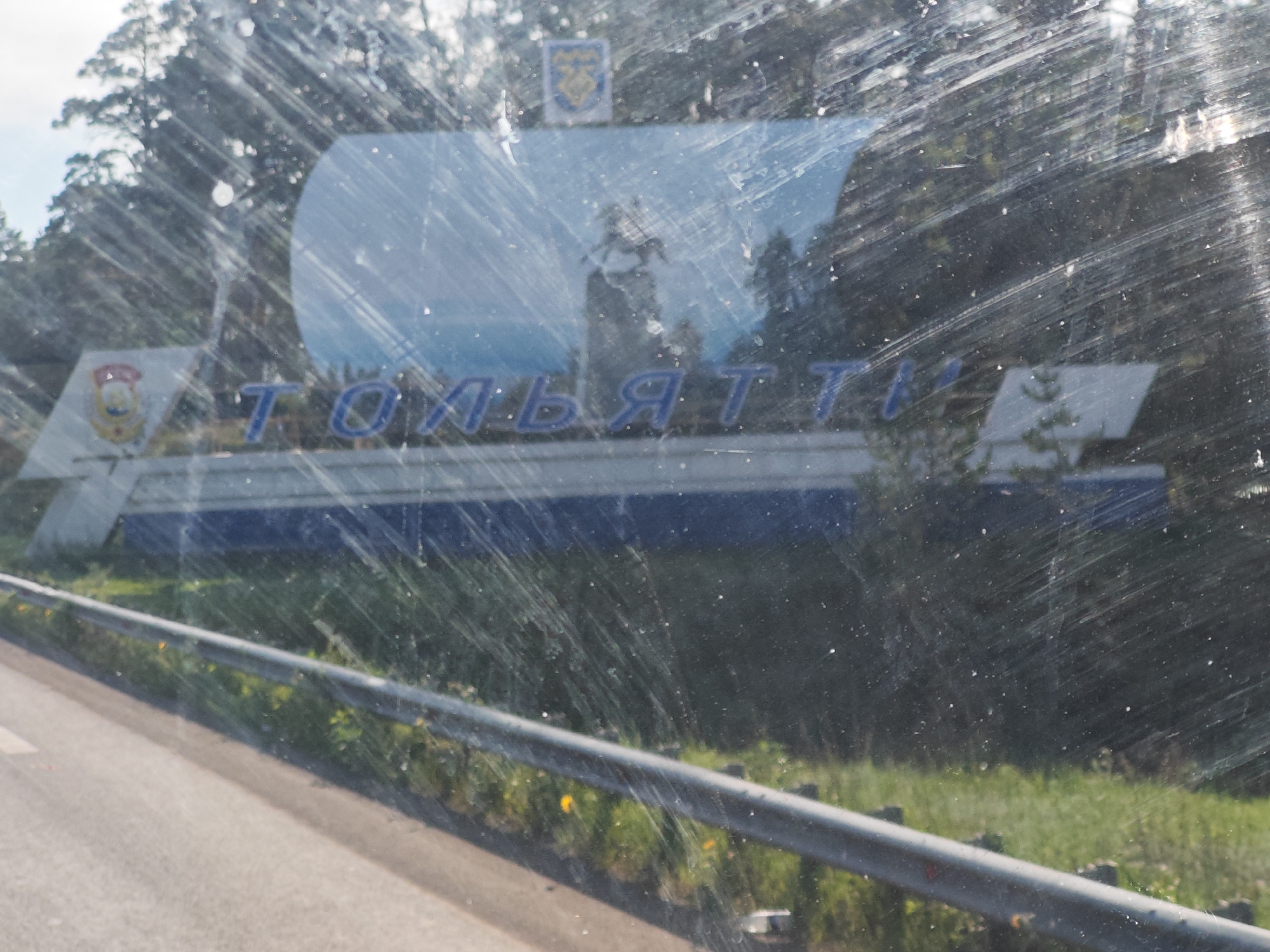 The image shows a sign in Russian with blue lettering that reads "ТОЛЬЯТТИ" (Tolyatti). The sign has a white and blue design and is partially obscured by reflections and smudges on a glass surface, likely a window. There are trees and greenery visible in the background, suggesting an outdoor location. The sign is positioned on the side of a road, with a guardrail visible in the foreground. The overall scene suggests the entrance to the city of Tolyatti, Russia.