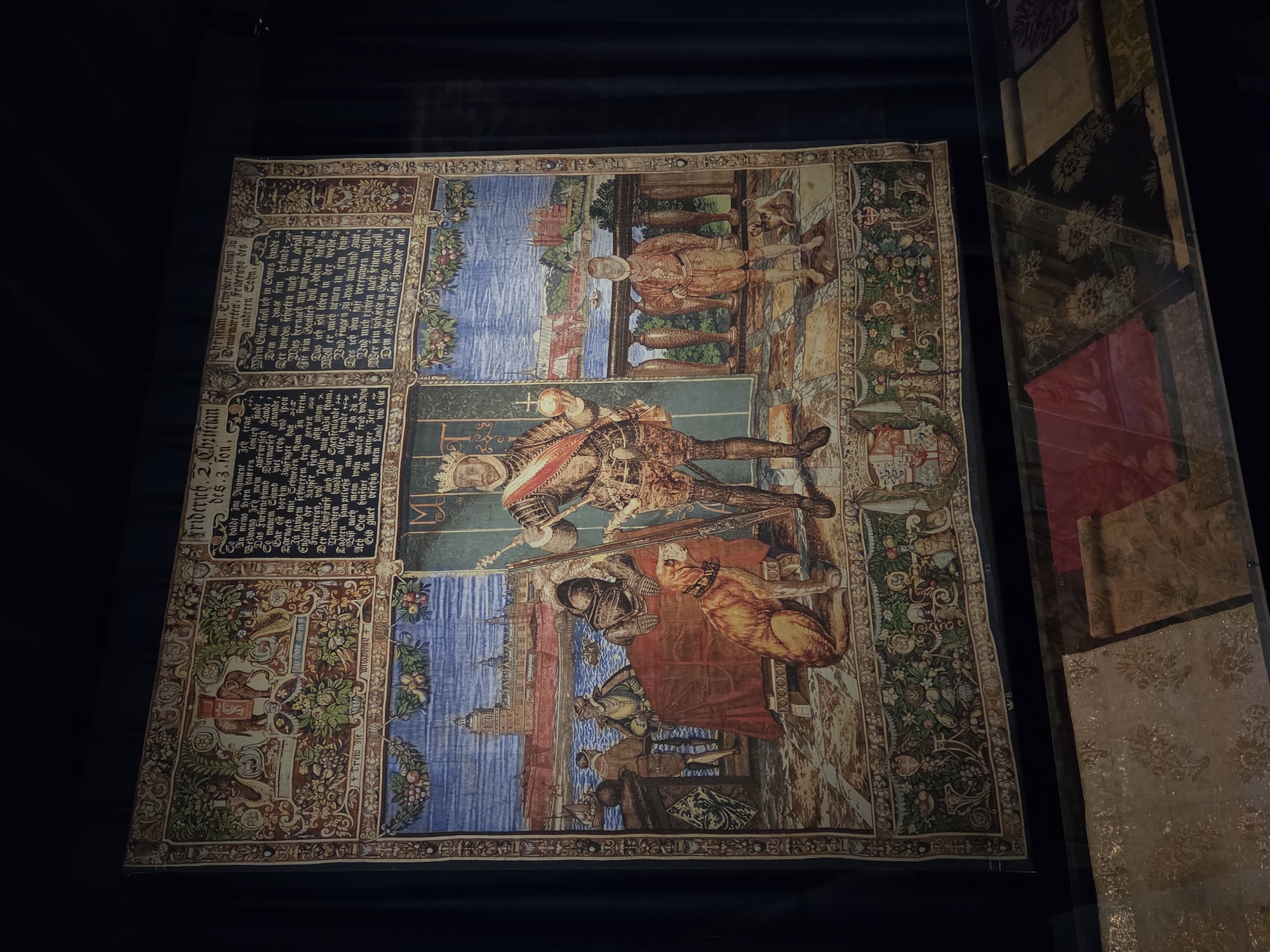 This image shows a tapestry on display in the city of Malmö, Sweden. The tapestry is elaborate and depicts a scene with several figures, including a king in armor holding a scepter and orb, a dog, and a young boy. There is also a depiction of a building with a waterway and boats in the background, and an elephant in a decorative border at the top. The tapestry appears to be framed and mounted on a wall, with a dark blue curtain behind it. Below the tapestry, there is a display case containing rolls of fabric with various patterns. The lighting in the scene suggests an indoor museum or exhibition setting. The text on the tapestry is in a historical script and is not fully legible.
