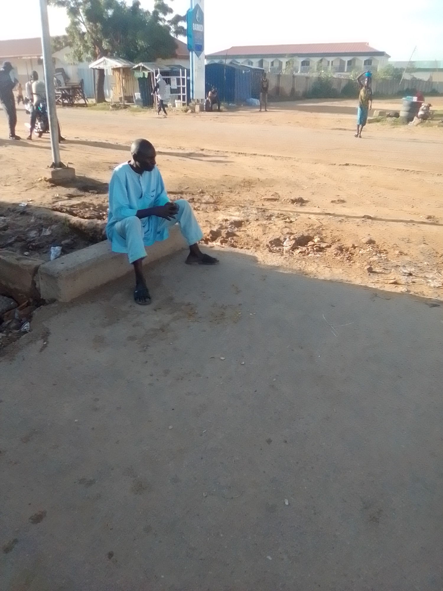 In Gombe, Nigeria, a man in a light blue traditional outfit sits on a concrete curb. He is facing towards the right of the frame, with his legs crossed. The man has dark skin and is bald. Behind him, a dusty road stretches out, lined with various structures including small buildings and what appears to be a gas station. In the background, there is a larger, light-colored building with arched windows. Several other people are visible in the distance, some walking and others standing. The environment appears to be an urban or semi-urban area with a dry, dusty landscape. The lighting suggests it is daytime.