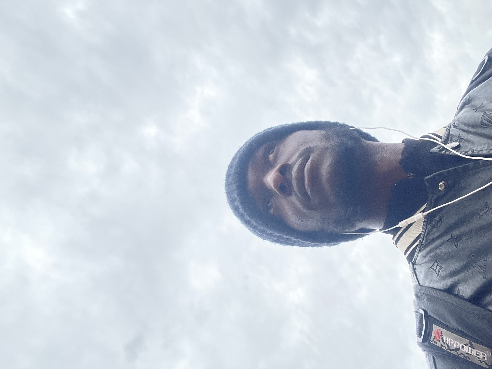 A low-angle shot captures a Black man wearing a dark blue beanie and a patterned jacket with white stripes on the collar. He is looking upwards. White earbuds are visible in his ears, with the cord trailing down his jacket. A black backpack with a red and white patch on the strap is on his left shoulder. The background is an overcast, cloudy sky. This image is not specific to Jos, Nigeria; it depicts a man outdoors under a cloudy sky.