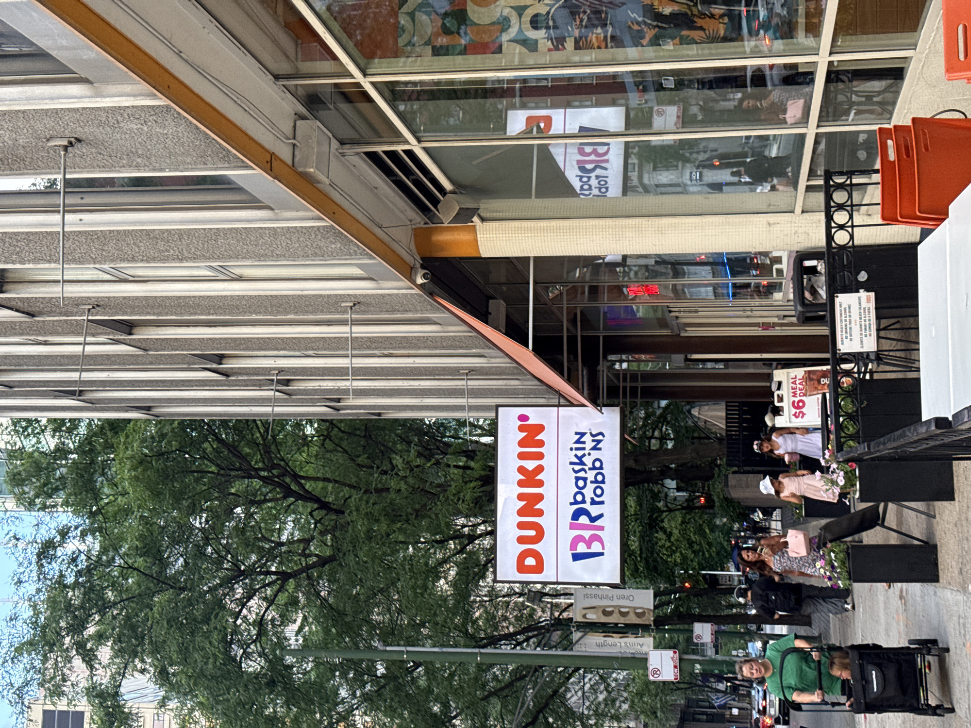 The image shows a street-level view of a Dunkin' and Baskin-Robbins combination store in an urban environment, likely Chicago, United States of America, based on the architecture and street signs. The store has a large, prominent sign featuring the Dunkin' logo in orange and the Baskin-Robbins logo in pink. The storefront features large glass windows and an awning. Outside the store, on the sidewalk, several people are visible. A woman in a green shirt pushes a stroller. Another woman in a pink dress is walking, holding a phone and a purse. Two young girls, one in a pink dress and the other in a white dress, are standing near a menu board displaying a "$6 Meal Deal". The street itself shows traffic and other pedestrians in the background. Lush green trees are present on the left side of the image, providing a contrast to the urban buildings. The overall scene depicts a busy commercial street with a focus on the food establishment and its customers.