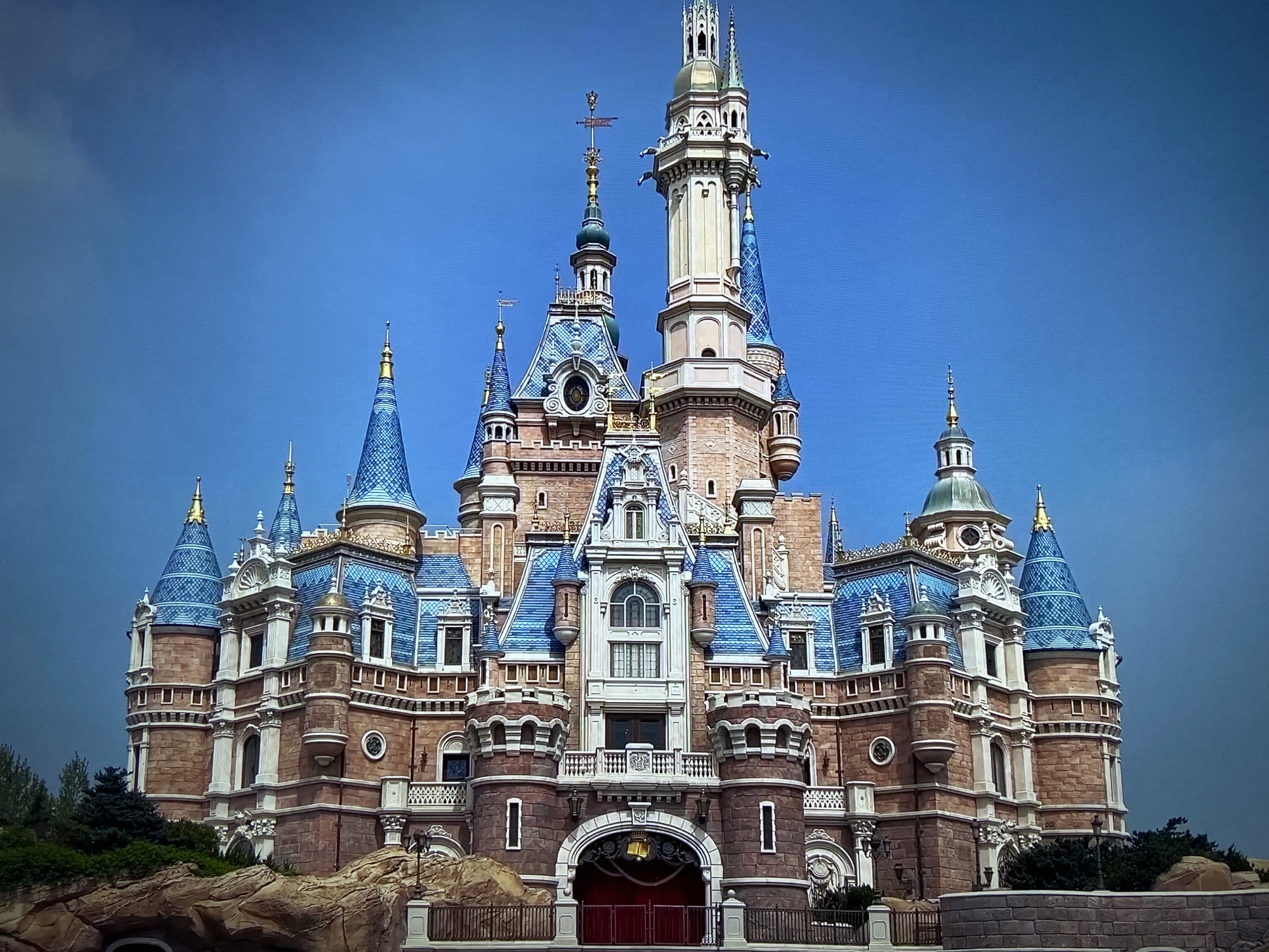 The image depicts a grand castle under a clear blue sky. The castle is constructed of a combination of brick and stone, featuring multiple towers and spires adorned with blue and gold detailing. The main entrance is marked by a large arched doorway with red doors. The building is situated on a rocky base with some greenery. The location is Unknown, Unknown.