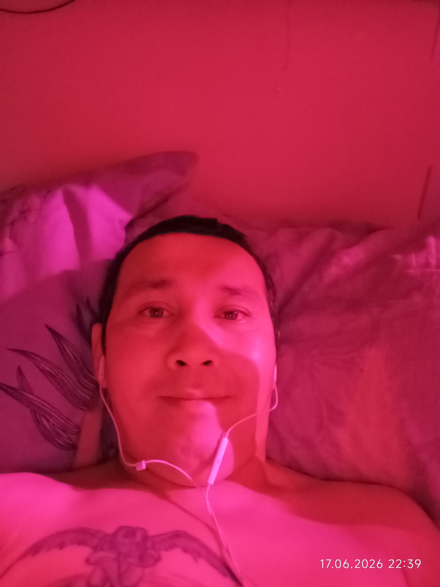 A close-up selfie shows a person lying down, illuminated by pink light. The person's head is angled slightly towards the camera, and they are wearing white earbuds. Their chest, partially visible, has a tattoo. The background appears to be a bed with patterned pillows. The image is dated "17.06.2026 22:39," suggesting it was taken in the evening. The setting is likely a bedroom in Nyurba, Russia.