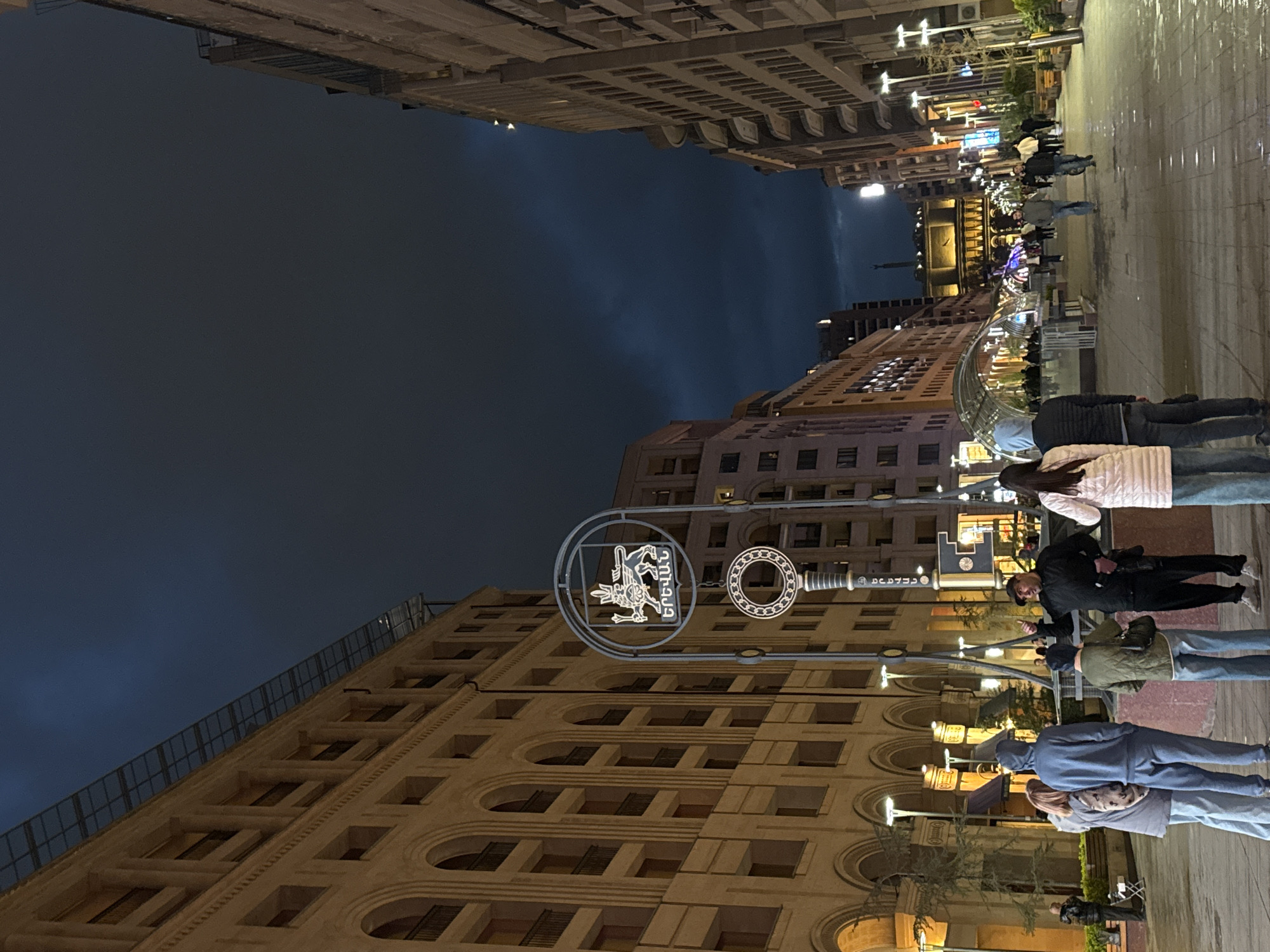 This nighttime urban street scene, set in Yerevan, Armenia, captures a vibrant atmosphere under a dark, overcast sky. The ground appears wet, reflecting the numerous artificial lights that illuminate the street and buildings.
Tall, multi-story buildings with intricate architectural details, including arched windows and textured facades, line both sides of the thoroughfare. The building on the left features a metal railing along its roofline. In the foreground, a prominent decorative street sign is visible, suspended over the street. It includes a circular frame enclosing a mythical creature and the Armenian text "ԵՐԵՎԱՆ" (Yerevan), alongside another key-shaped element.
Several people are seen walking on the right side of the street, dressed in casual clothing such as jackets and jeans. Some walk individually, while others move in small groups, suggesting an evening stroll or daily activity. Their faces are not clearly discernible. In the background, further down the street, an impressive, brightly lit building or monument can be seen, possibly incorporating illuminated water features or fountains, indicating a significant landmark within the city. The overall scene depicts a lively but calm urban evening.