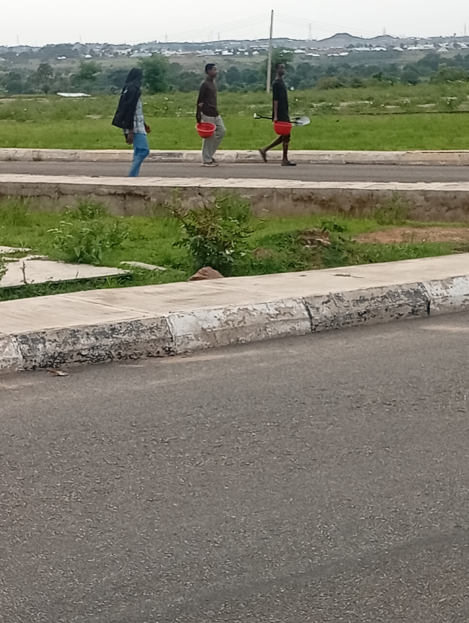 Three individuals are walking on a paved road beside a grassy area, with a distant background of rolling hills and scattered buildings under an overcast sky. The person on the left, wearing a black jacket over a checkered shirt and blue pants, carries a large black bag slung over their shoulder. The person in the middle, dressed in a brown long-sleeved shirt and light-colored pants, holds a red bucket in their right hand. The person on the right, in a black t-shirt and dark shorts, also carries a red bucket in their right hand, and a shovel is resting on their right shoulder, with its blade inside the bucket. A white concrete curb separates the road from the grassy area. The overall scene appears to be within the city of Yelwa, Nigeria.