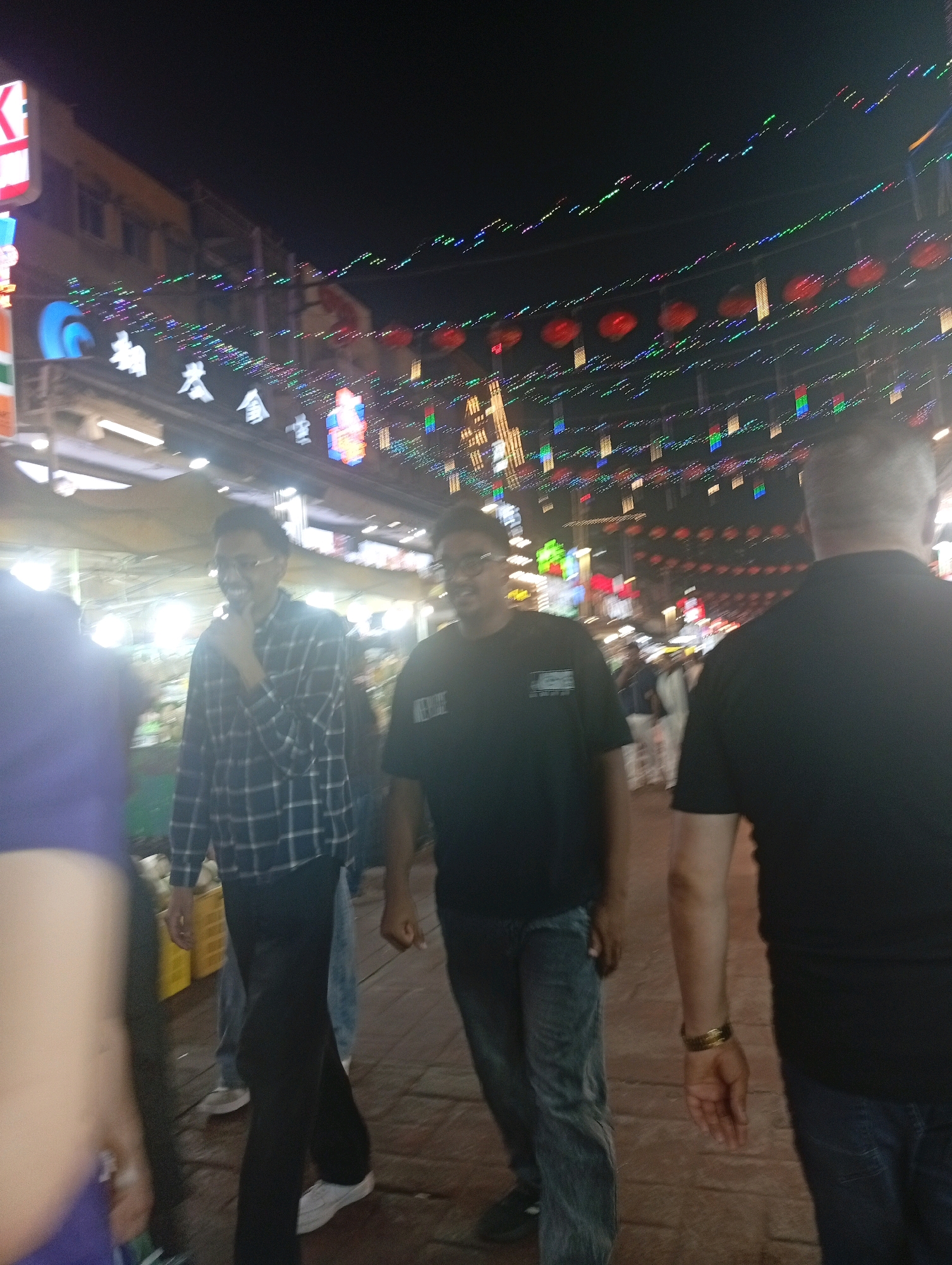 The image depicts a nighttime scene on a street in Kuala Lumpur, Malaysia, likely a tourist area such as Petaling Street, characterized by festive decorations. Several individuals are visible, some in focus and others blurred due to motion or camera focus.
In the foreground, a person wearing a purple garment on their left side is partially visible, appearing out of focus. To their right, a man in a blue and white checkered shirt and dark pants is in focus, smiling and touching his chin. He is wearing glasses. Next to him, a man in a black t-shirt and blue jeans is also in focus, walking. Another man in a black t-shirt is seen from the back, also in focus, wearing a watch on his left wrist.
The background is a blur of lights and structures, suggesting a bustling street environment. Strings of colorful lights and red lanterns are strung across the street, illuminating the night. Buildings with signs are visible, some with Chinese characters, typical of Chinatown areas. The ground appears to be paved with bricks. The overall atmosphere is that of a lively night market or shopping street.