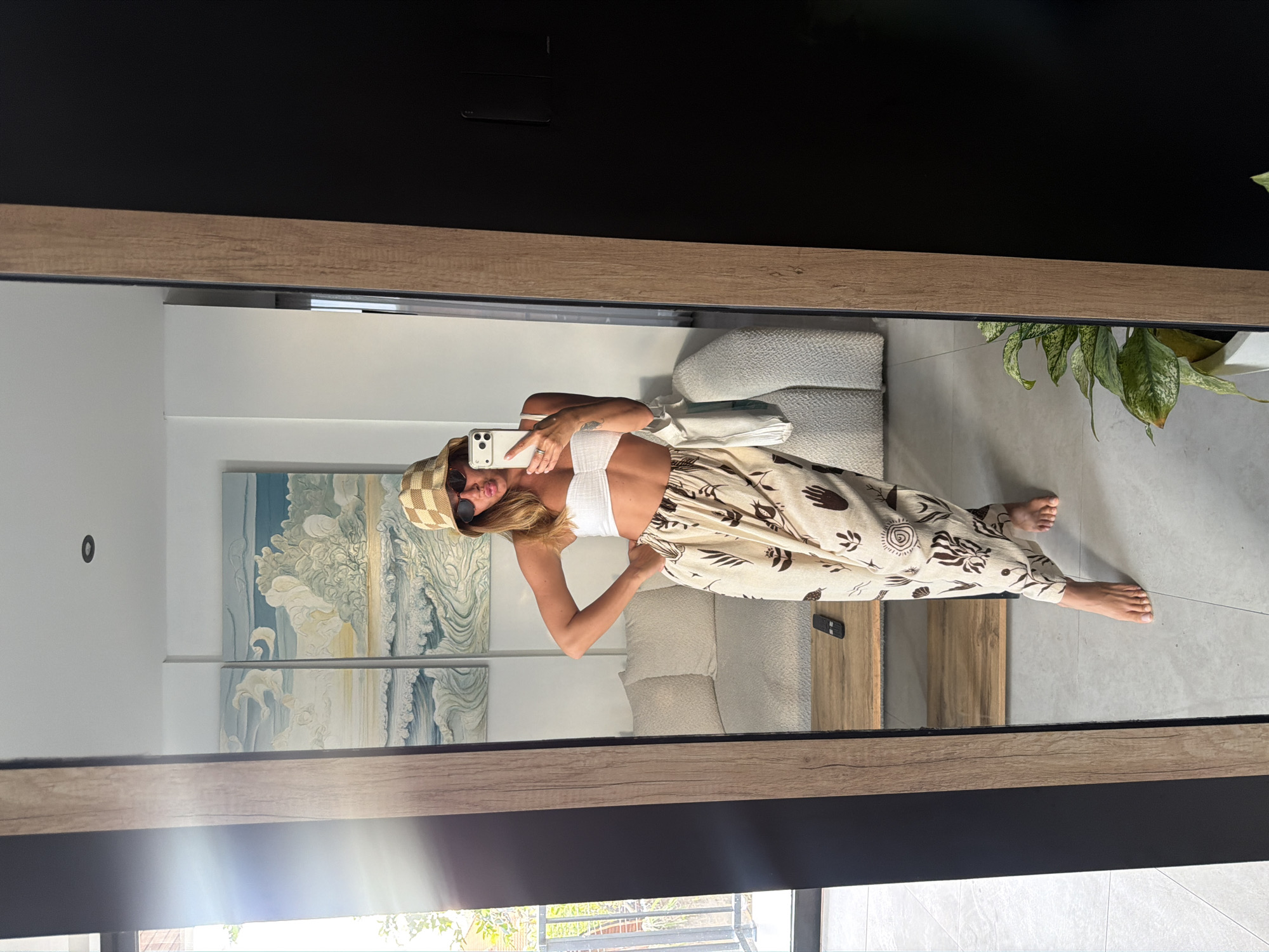 A woman is taking a mirror selfie in a room with a sofa, two paintings, and a plant. She is wearing a white bandeau top, patterned wide-leg pants, a checkered bucket hat, and sunglasses. Her left hand is on her hip, and she is holding her phone in her right hand. The room has white walls and a light-colored tiled floor. There is a wooden framed mirror and a wooden coffee table with a remote control on it. The paintings appear to be of a beach scene with waves and clouds.
