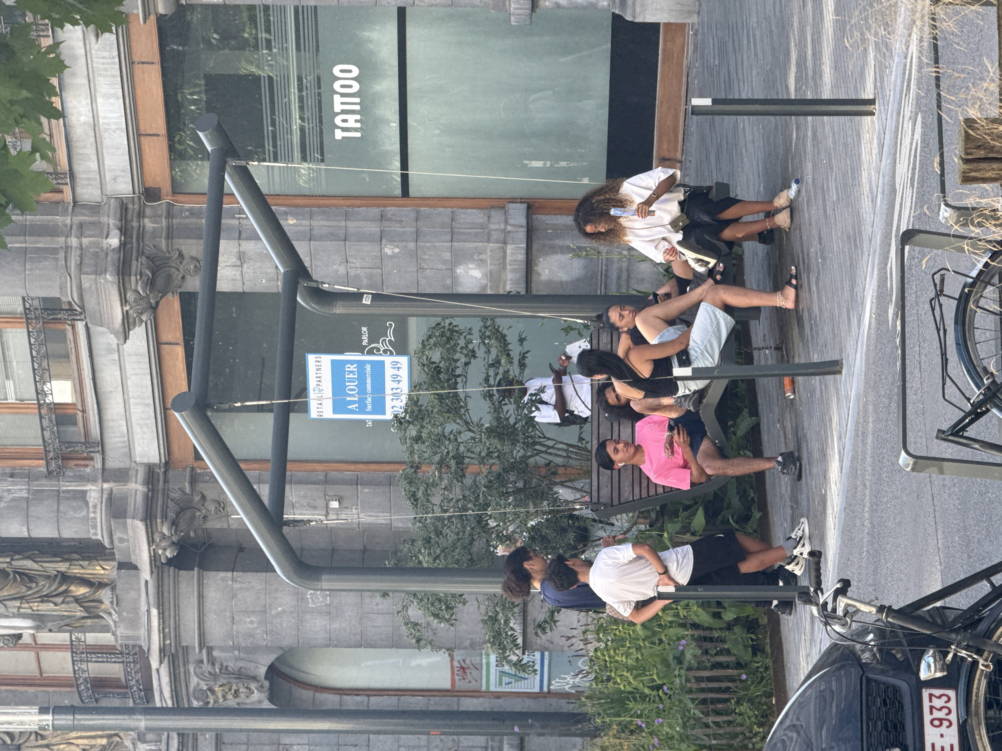 The image depicts a street scene in Brussels, Belgium, with several people sitting on a bench. The setting appears to be outdoors, possibly in a commercial area given the presence of a building with "TATTOO PARLOR" signage and a "A LOUER" (For Rent) sign on the facade. The time of day seems to be daytime, with bright lighting suggesting fair weather.
The individuals on the bench are casually dressed, and their postures suggest relaxation. One person, a woman with curly hair, is holding a bottle. Another person is wearing a pink t-shirt and dark shorts, while others are in white t-shirts and dark shorts or skirts. There's a bicycle parked nearby, a common sight in urban European settings. The architecture of the building in the background shows ornate details, hinting at an older part of the city.
The visible text includes "TATTOO PARLOR", indicating the nature of a business located there. The "A LOUER" sign suggests a commercial space is available for lease. Additionally, a phone number "02 303 49 49" is visible on the rental sign. The license plate of a car in the foreground shows "E-933". The overall mood seems relaxed and ordinary, capturing a moment of everyday life in the city.