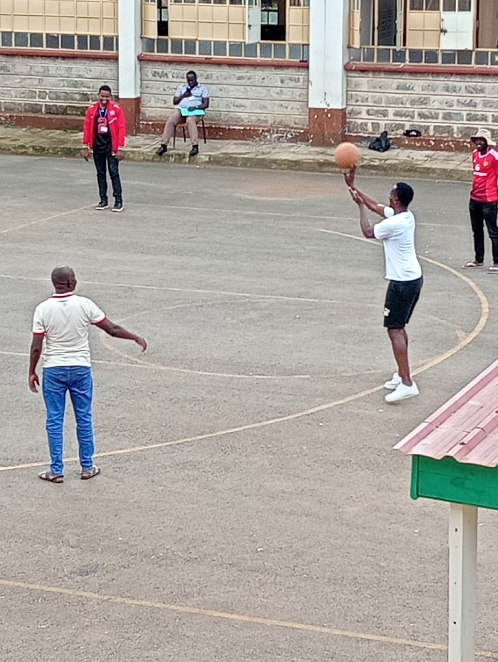 An outdoor scene captured in Riara Ridge, Kenya, depicts five men on a paved court with faded white and yellow lines, resembling a basketball or multi-purpose court. In the foreground to the right, a man in a white t-shirt, black shorts, and white sneakers is captured mid-action, holding a basketball in a shooting form, poised to release it. His gaze is directed towards an unseen hoop.
To the left, with his back partially turned to the camera, another man in a white t-shirt, blue jeans, and sandals stands observing, his left arm extended slightly. Further back on the left side of the court, a man wearing a red jacket, black trousers, and black shoes stands smiling, looking towards the center of the activity. Behind him, another man sits on a chair against a low stone wall, wearing a light blue collared shirt and dark trousers, holding what appears to be a clipboard or papers. His posture suggests he might be an official or observer. On the far right, partially visible, a fifth man in a red t-shirt and dark trousers, wearing sandals, stands near the edge of the court.
The background features a light-colored stone building with multiple windows and a reddish-brown brick pattern along the top of the wall. A green and red-roofed structure is visible in the lower right foreground. The lighting suggests it is daytime, possibly overcast, creating soft, even illumination. No visible text is discernible on clothing or surrounding structures. The overall atmosphere appears casual, possibly a practice session or friendly game.