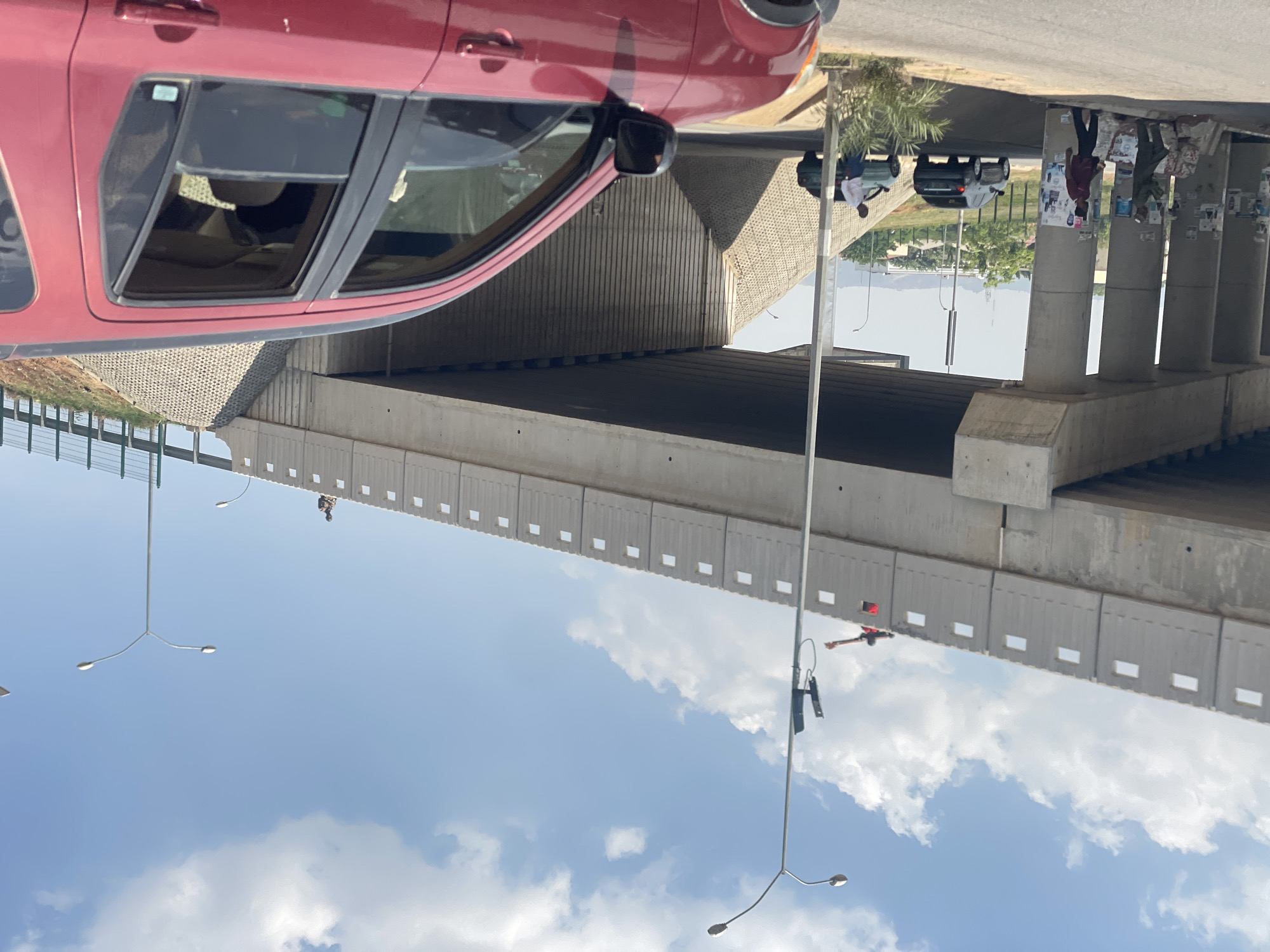 This daytime image captures a multi-level urban transportation scene, likely in Abuja, Nigeria, characterized by an overpass and busy roadways under a clear sky.
In the foreground, on the left, a significant portion of a red car, possibly an SUV or crossover, is prominently featured. Its rear passenger windows are partially rolled down, revealing glimpses of a light-colored interior. The car appears to be stationary on a road that runs parallel to the main thoroughfare.
The central and dominant feature of the image is a massive concrete overpass or flyover. A road passes underneath this structure, forming an underpass, while another elevated road runs along its top. The side barriers of the overpass are made of concrete panels with a repeating pattern of small rectangular openings.
Several individuals and vehicles are visible across the scene. On the upper road, above the overpass structure, two dark-colored cars are visible, along with a person in a white shirt standing near the left-most dark car. Further to the right, several concrete pillars support the overpass, some adorned with various posters or advertisements. A person in a dark top is seen standing near these pillars. On the elevated road of the overpass, a person wearing a red top and dark trousers is seen walking near the concrete barrier on the right side.
The sky is a vibrant blue with scattered, fluffy white clouds, indicating sunny and pleasant weather conditions. Multiple streetlights are strategically placed throughout the urban landscape, with one notably long pole extending from the lower left towards the upper right. The scene conveys a sense of active urban life and infrastructure. No discernible text is clearly readable in the image.