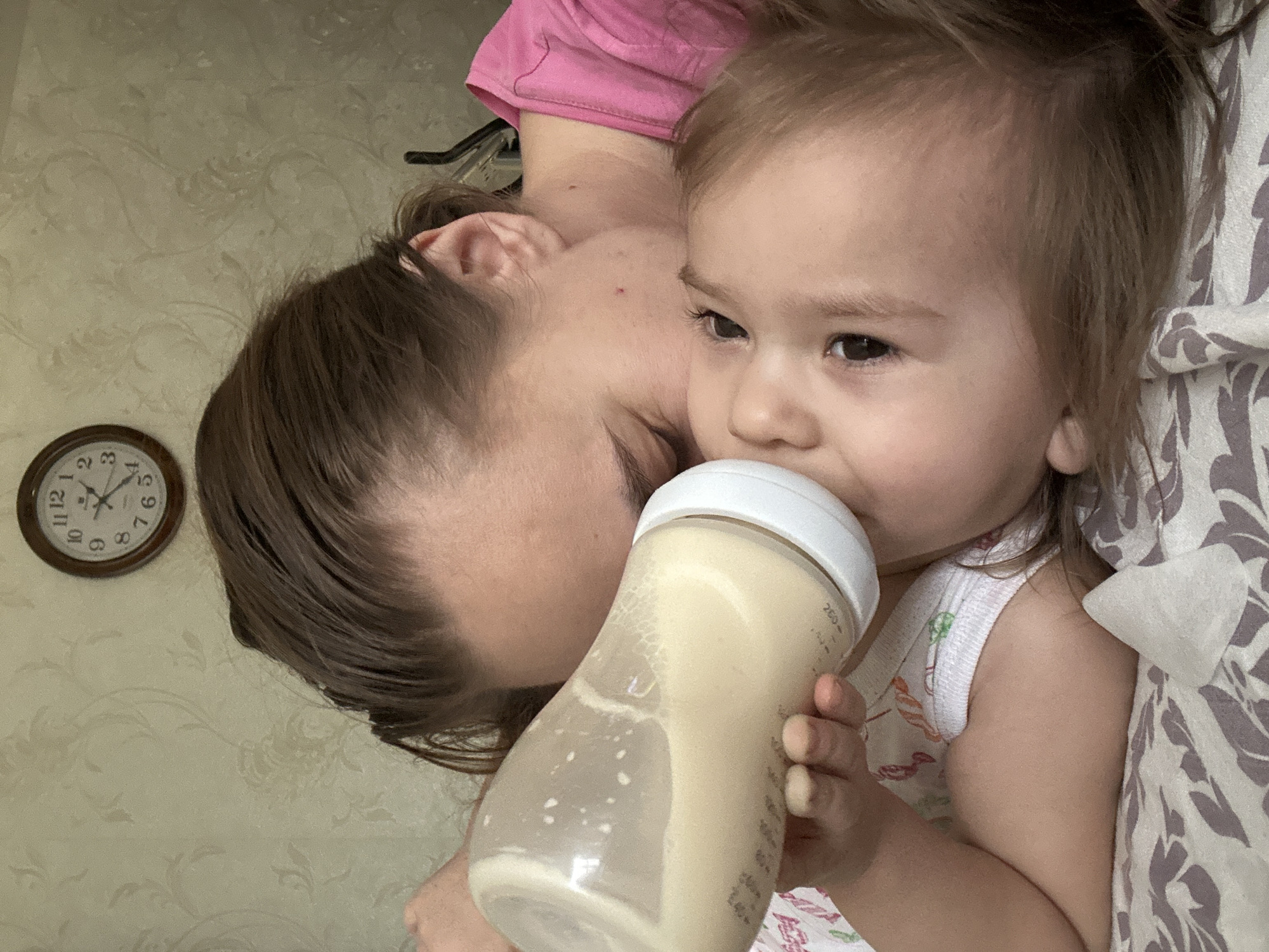 This image presents a heartwarming, overhead view of an adult and a young child in an intimate moment, likely in a home setting in Uva, Russia. The adult, whose face is largely obscured but shows parts of a cheek and ear with brown hair, is wearing a pink top and is positioned closely beside the child.
The child, a toddler or young baby with light brown hair and dark eyes, is the central focus, actively drinking from a clear baby bottle with a white cap. The bottle contains a light-colored liquid, possibly formula or milk, with visible bubbles indicating ongoing consumption. "200 ml" is clearly visible as a measurement mark on the side of the bottle. The child is dressed in a light-colored garment and is nestled against a patterned fabric, featuring a grey and purple floral design, possibly a blanket or bedsheet.
In the background, a round wall clock with a wooden frame is visible, indicating the time is approximately 1:40 (minute hand at 8, hour hand between 1 and 2). The wall behind them is adorned with light beige wallpaper featuring a subtle, embossed floral or swirl pattern. The overall atmosphere is one of comfort, closeness, and gentle care, capturing a quiet feeding moment between the parent and child.