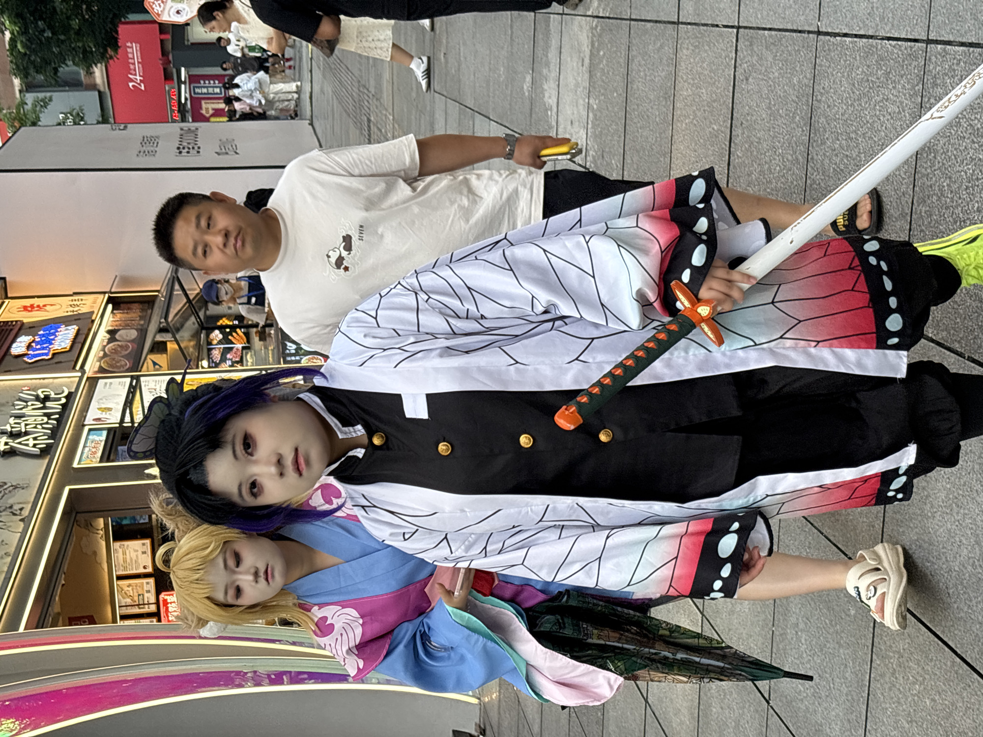 In Yuzhong District, China, on a paved city sidewalk, two individuals dressed in anime-inspired cosplay are walking. The person in the foreground is dressed as Shinobu Kocho from Demon Slayer, complete with a black uniform, a white haori with a butterfly wing pattern, and a prop katana. Their hair is styled in a dark bun with purple streaks. To their left and slightly behind is another cosplayer with blonde hair, wearing a blue and pink kimono-like garment and holding a patterned umbrella. They are walking away from the camera. Behind them, an Asian man in a white t-shirt with a panda graphic is walking towards the camera, looking ahead. The background shows a street with illuminated shop fronts, neon signs in Chinese characters, and other pedestrians. The lighting suggests it is daytime.