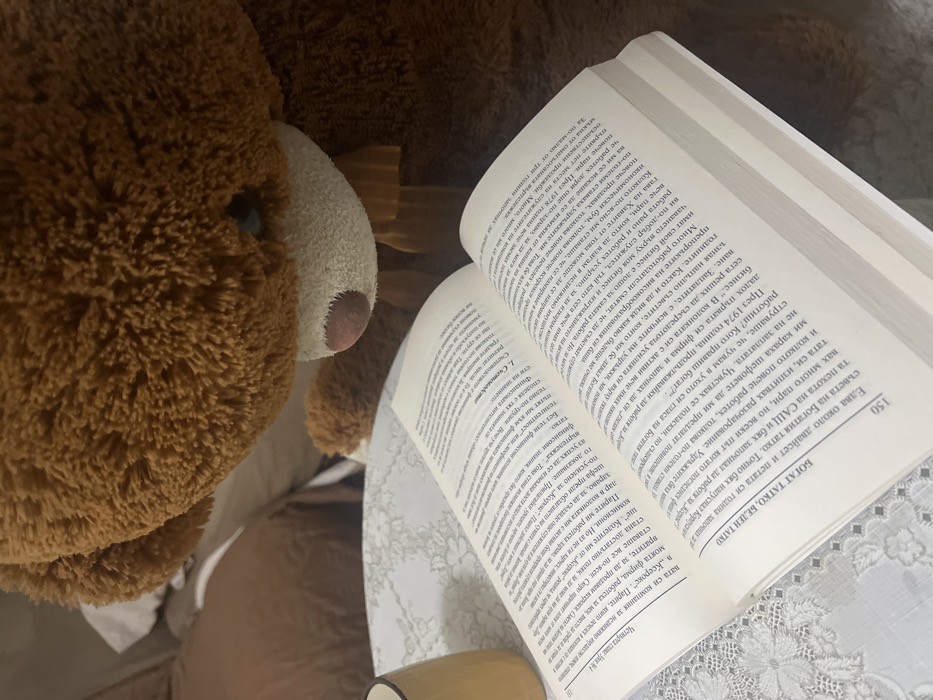 A large, shaggy brown teddy bear with a lighter snout and a brown bow sits positioned over an open book. The book displays white pages with black Cyrillic text, including the heading "БОГАТ ТАТКО, БЕДЕН ТАТКО" and page number "150". It rests on a white surface with a subtle floral or lace pattern, alongside a partially visible beige ceramic mug positioned to the left. The background shows blurred brown and white soft furnishings, suggesting an indoor domestic setting in Sedlartsi, Bulgaria.