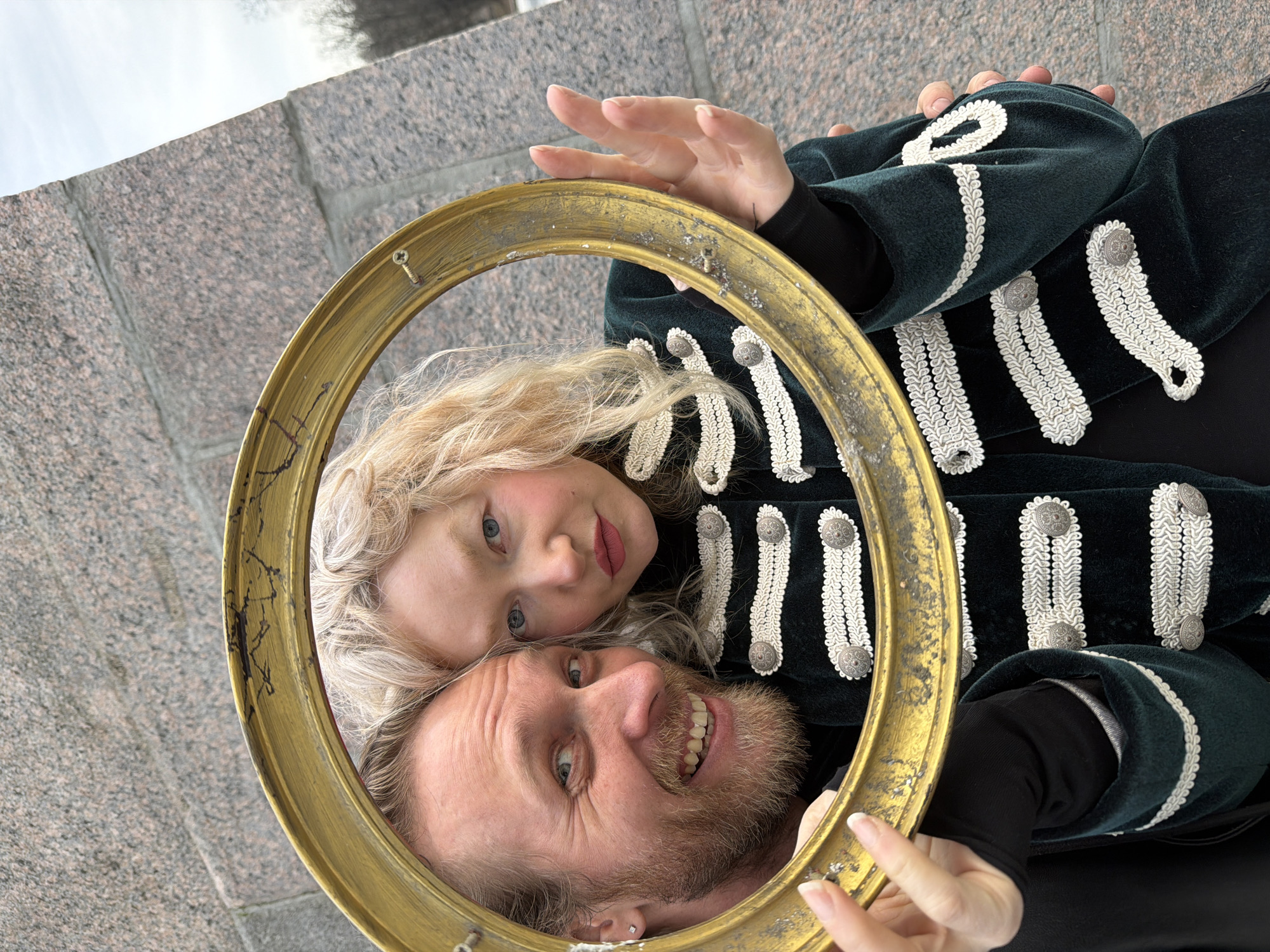 A man and a girl, probably father and daughter, are photographed holding a gold-colored round frame. The man is at the bottom with his head upwards, grinning broadly. The girl is above him, smiling softly. Both are inside the frame. The man has a beard, fair hair, and an earring in his left ear. The girl has fair, curly hair and blue eyes. The person holding the frame is wearing a dark green jacket with decorative white braid and buttons, and a black long-sleeved shirt underneath. The background shows a stone wall and overcast sky. Based on the wall and general aesthetic, the image was likely taken in Saint Petersburg, Russia.