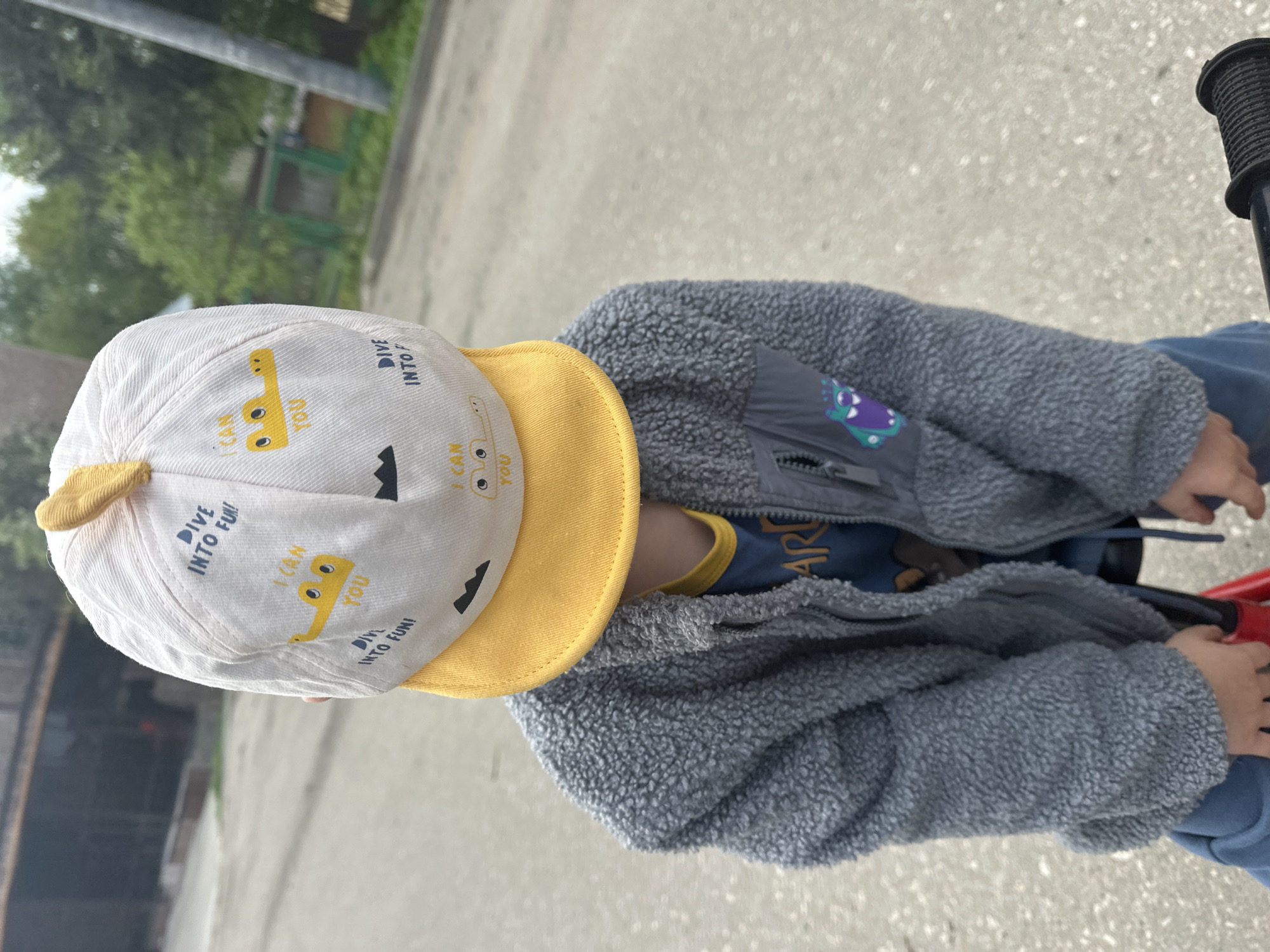 A toddler is riding a bicycle on a paved surface. The toddler is wearing a white and yellow baseball cap with the phrase "DIVE INTO FUN!" and a picture of a yellow submarine. The toddler is also wearing a gray fleece jacket and blue pants. The bicycle has black handlebars and a red frame. The background shows a road and some greenery, suggesting the scene is outdoors, possibly a park or residential area. The exact location in Nizhny Novgorod, Russia, cannot be determined from the image.
