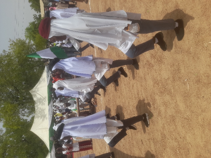 The image shows a group of people, presumably students, performing a synchronized dance routine in a field. They are wearing white robes and black pants and are all facing the right side of the image. The students are in a line formation, and they are all stepping forward with their left feet, their right foot raised behind them. Their hands are raised, palms facing inward, and they are wearing white gloves. There is a Nigerian flag on a pole in the background. There is a large white tent in the background, which is likely a canopy for an event. The image was taken in an outdoor setting, with a sandy ground. This location is at the coordinate (12.187, 6.65) in Gusau, Nigeria.