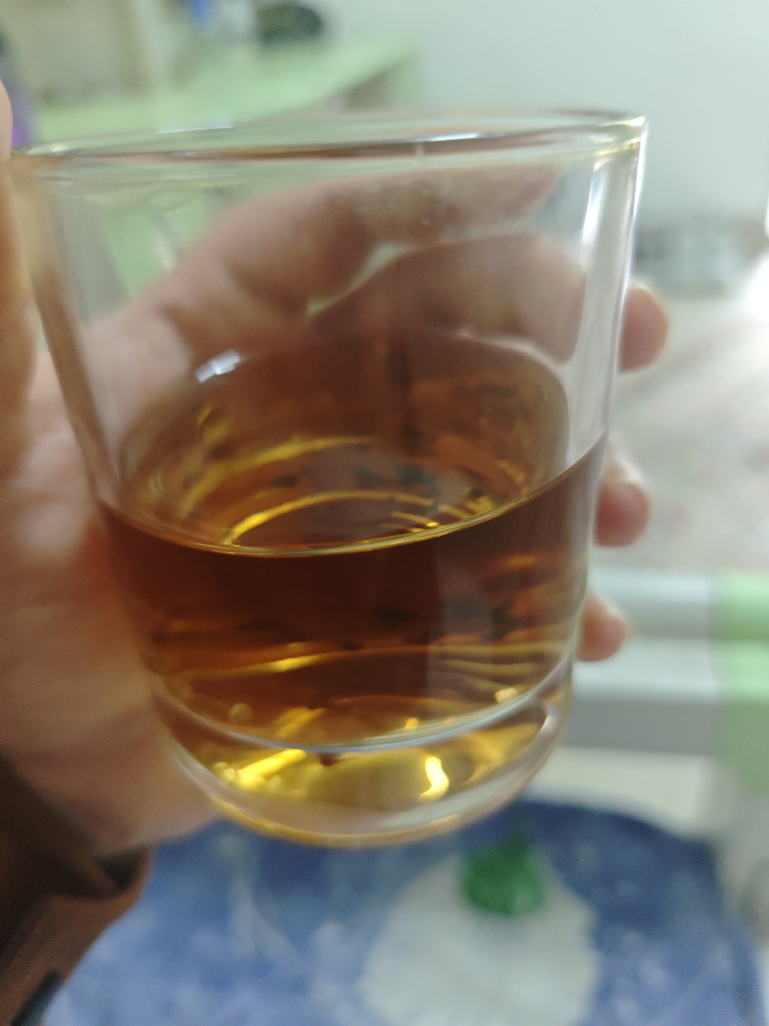 A hand holds a clear glass approximately half-full of a dark amber liquid. The glass is cylindrical with a thick base. The liquid's surface is reflective. The background is blurred, but visible elements include a green-topped table, a blue-and-white patterned surface, and a white object. The photograph may have been taken in Chongqing High-tech Zone, China. No people are entirely visible besides the hand holding the glass. There are no visible actions or interactions beyond the hand holding the glass. The environment is domestic.