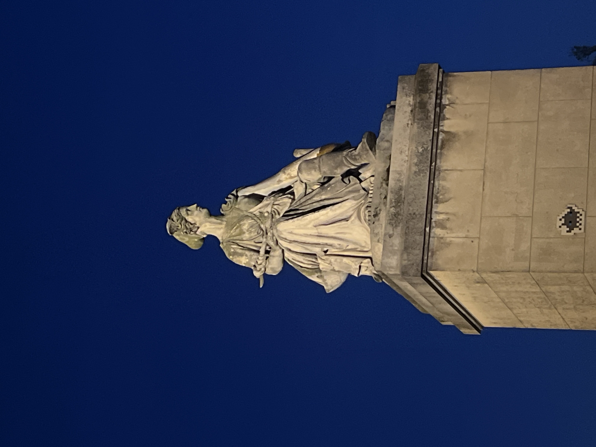 The image shows a stone statue of a seated female figure against a dark blue sky. The statue is positioned atop a tall, rectangular stone pedestal. The female figure appears to be wearing classical drapery, with her hair styled in an updo. She is holding a slender object, possibly a scroll or a baton, in her hands. The statue and pedestal are lit from below, casting a subtle glow on the stone.
This statue is likely located in Paris, France, specifically on the Pont Alexandre III. The Pont Alexandre III is a highly ornate bridge spanning the Seine River, known for its elaborate sculptures and decorative elements, including statues of allegorical figures.