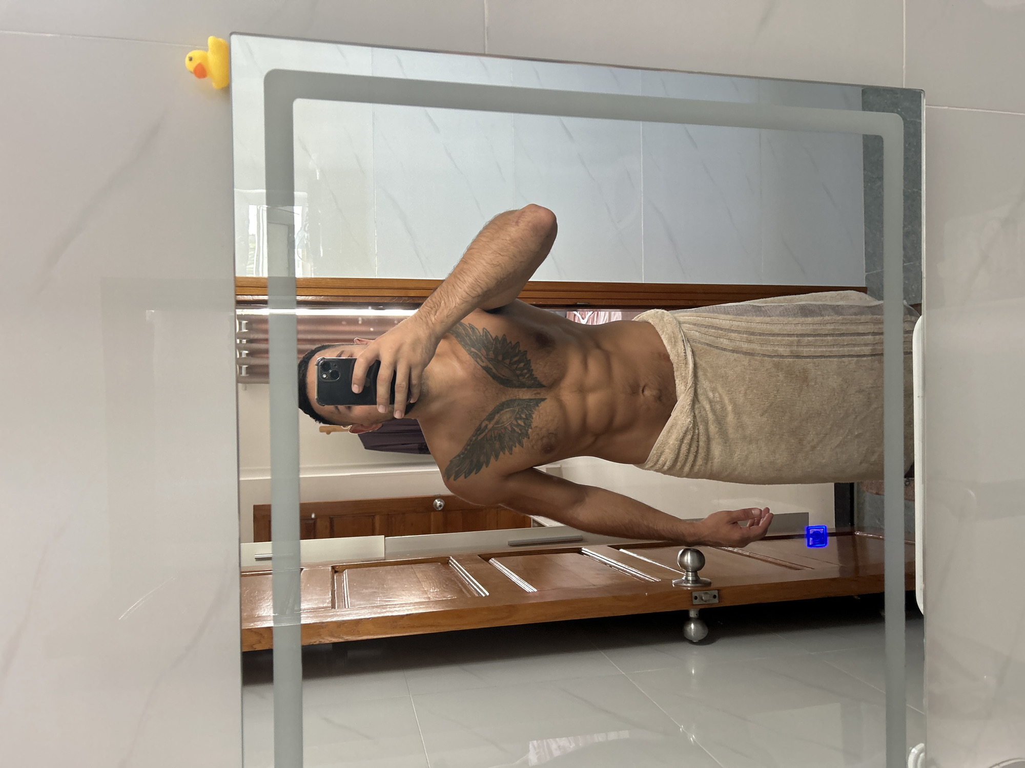 A shirtless man with a towel wrapped around his waist and an angel wing tattoo on his chest stands in front of a mirror in a bathroom. He is holding a smartphone and taking a selfie. The bathroom features white tiled walls, a wooden door, and a rubber duck on top of the mirror. The scene appears to be in Nha Trang Ward, Vietnam, based on the metadata.