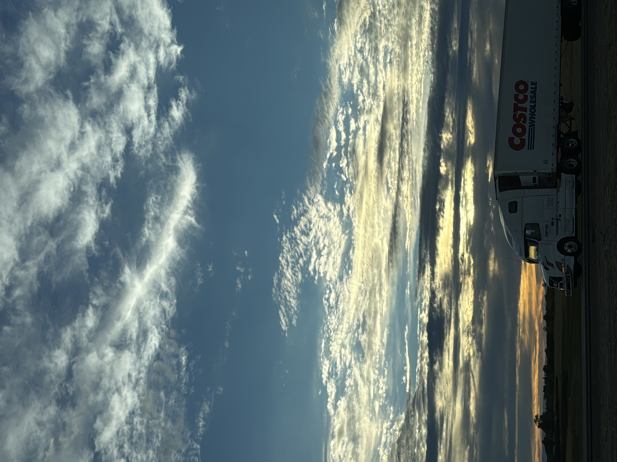 The image shows a Costco semi-trailer truck driving on a highway in Elk Grove, United States of America. The truck is white with the Costco Wholesale logo on the side. The sky is a dramatic mix of blue, grey, and white clouds, with hints of sunset colors on the horizon. The truck is in the foreground, and the background consists of a flat, open landscape with some trees. The overall impression is one of a journey or movement, with the vastness of the sky contrasting with the man-made object of the truck.