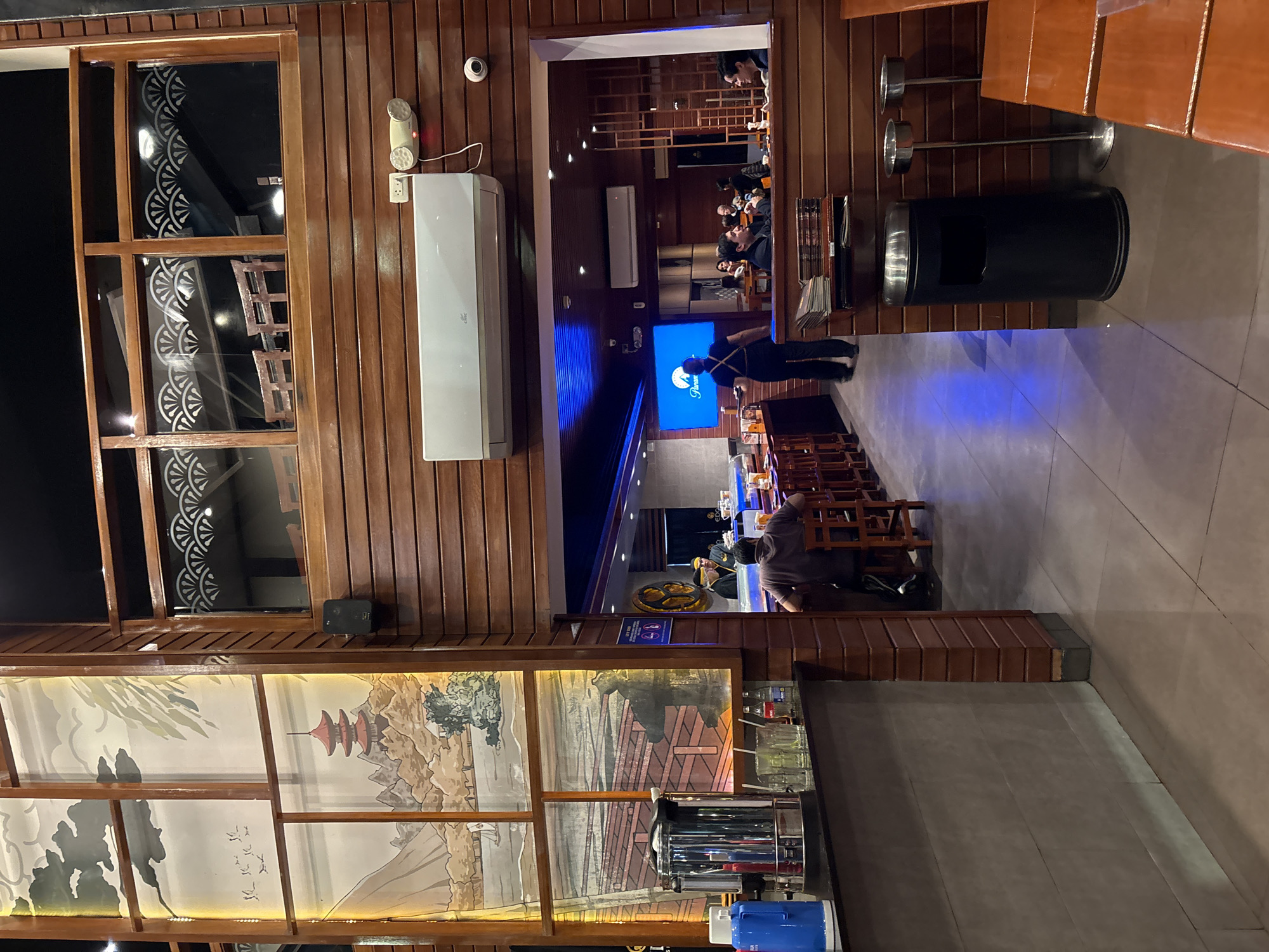 The image depicts the interior of a Japanese restaurant, likely in Magdalena del Mar, Peru, characterized by its extensive use of dark wood paneling and ambient lighting. In the foreground, a bar area is visible with a stainless steel coffee maker, a blue thermos, glasses, and bottles on the counter. To the right, a man is seated at the bar, facing away from the camera. Beyond the bar, an open entryway reveals the main dining area. Here, a staff member in dark attire is walking past a sushi bar where patrons are seated. A large screen displays the "Paramount" logo. The restaurant features tiered seating and decorative elements reminiscent of Japanese art, such as paintings of landscapes and pagodas on the upper levels. The overall atmosphere appears to be upscale and contemporary, with a clear emphasis on a Japanese theme.