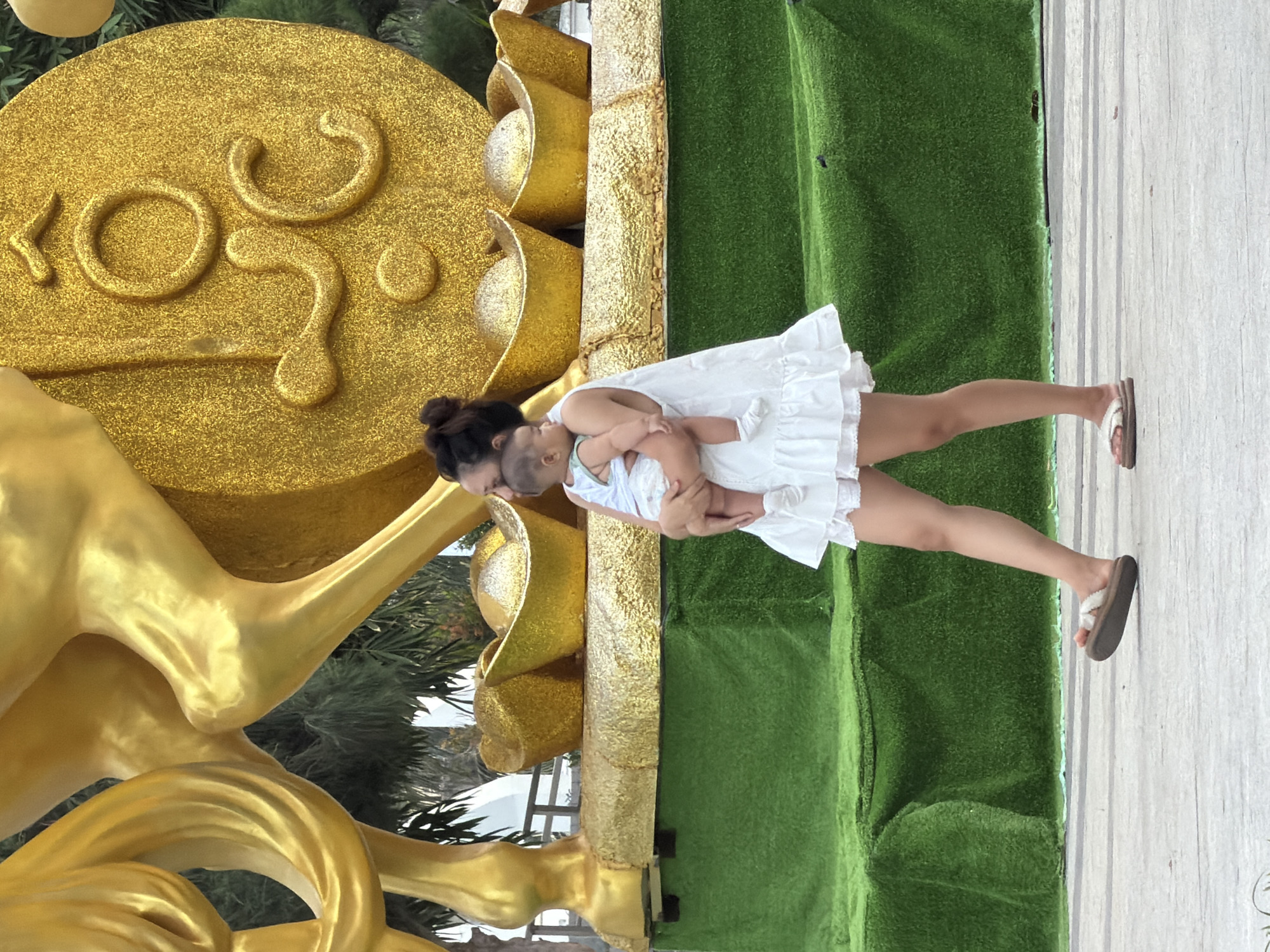 A mother carries her baby while walking in front of a large gold sculpture in Nha Trang, Vietnam. The sculpture appears to be an abstract representation of a horse and coins. The mother wears a white dress and flip-flops, and the baby is dressed in a white onesie. The background features lush green artificial turf and palm trees, suggesting an outdoor recreational area or a decorative display. The overall scene is bright and sunny, indicating daytime.