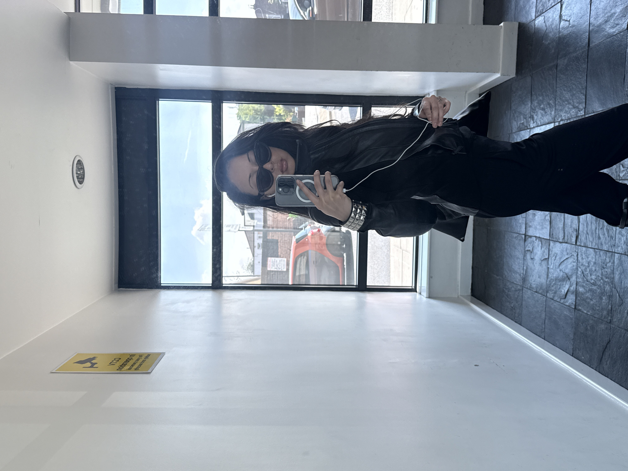 A woman is taking a mirror selfie in a modern building in London. She is wearing a black leather jacket, black pants, and sunglasses. Her dark hair is long and loose. She is holding a smartphone with a clear case, and there is a white cable attached to it. She is positioned between a window and a wall. Through the window, the cityscape is visible. On the wall is a sign. The floor has a glossy white finish, contrasting with a tiled area to the right. The building's interior is well-lit.