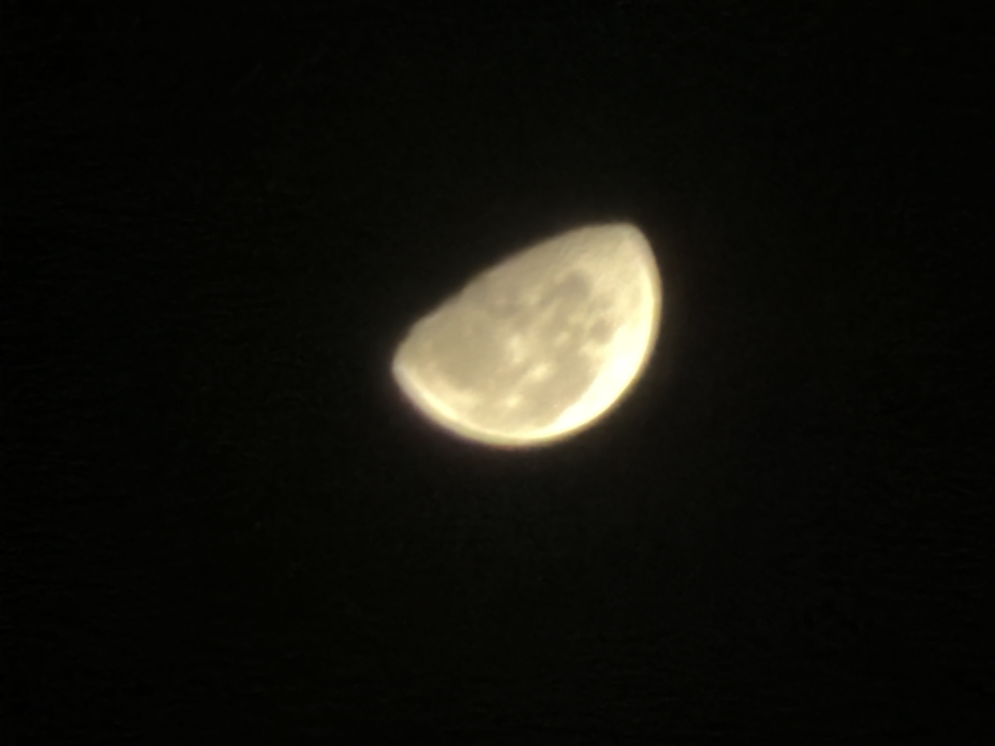The image displays a celestial body, identifiable as the Moon, captured against a uniformly dark, black background, indicative of a nighttime sky. The Moon is illuminated, appearing in a waxing gibbous phase, with approximately two-thirds of its disk visible and brightly lit. Its color is a warm yellow-gold. Visible features on the lunar surface are indistinct due to a slight blur or soft focus, but darker areas, likely maria, are discernible within the illuminated portion. The edges of the Moon show a subtle glow or chromatic aberration. There are no other celestial objects, atmospheric phenomena, or terrestrial elements visible in the frame. The image does not contain any people, subjects, objects, or environmental details that would indicate a specific terrestrial location, including the city of New Delhi, India.