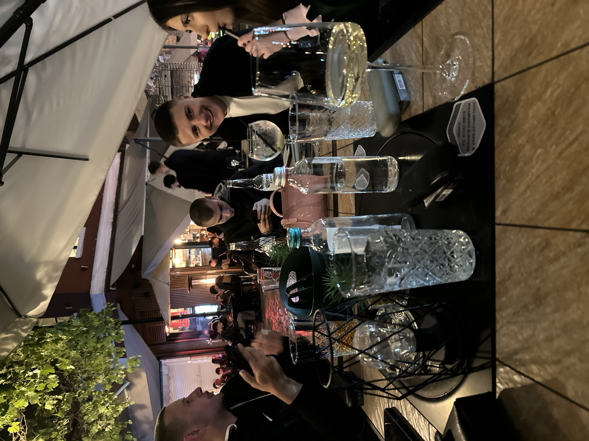 The image shows a group of people dining outdoors at a restaurant. Two people are visible at the top, one smiling at the camera. The setting is likely a restaurant patio with umbrellas providing shade.
On the table, there are various drinks, including cocktails, water bottles, and glasses. One of the bottles appears to be a Corona Extra.
The location is the city of Plovdiv, Bulgaria. The restaurant patio appears to be well-lit with lights strung overhead. Several people are seated further back at other tables.